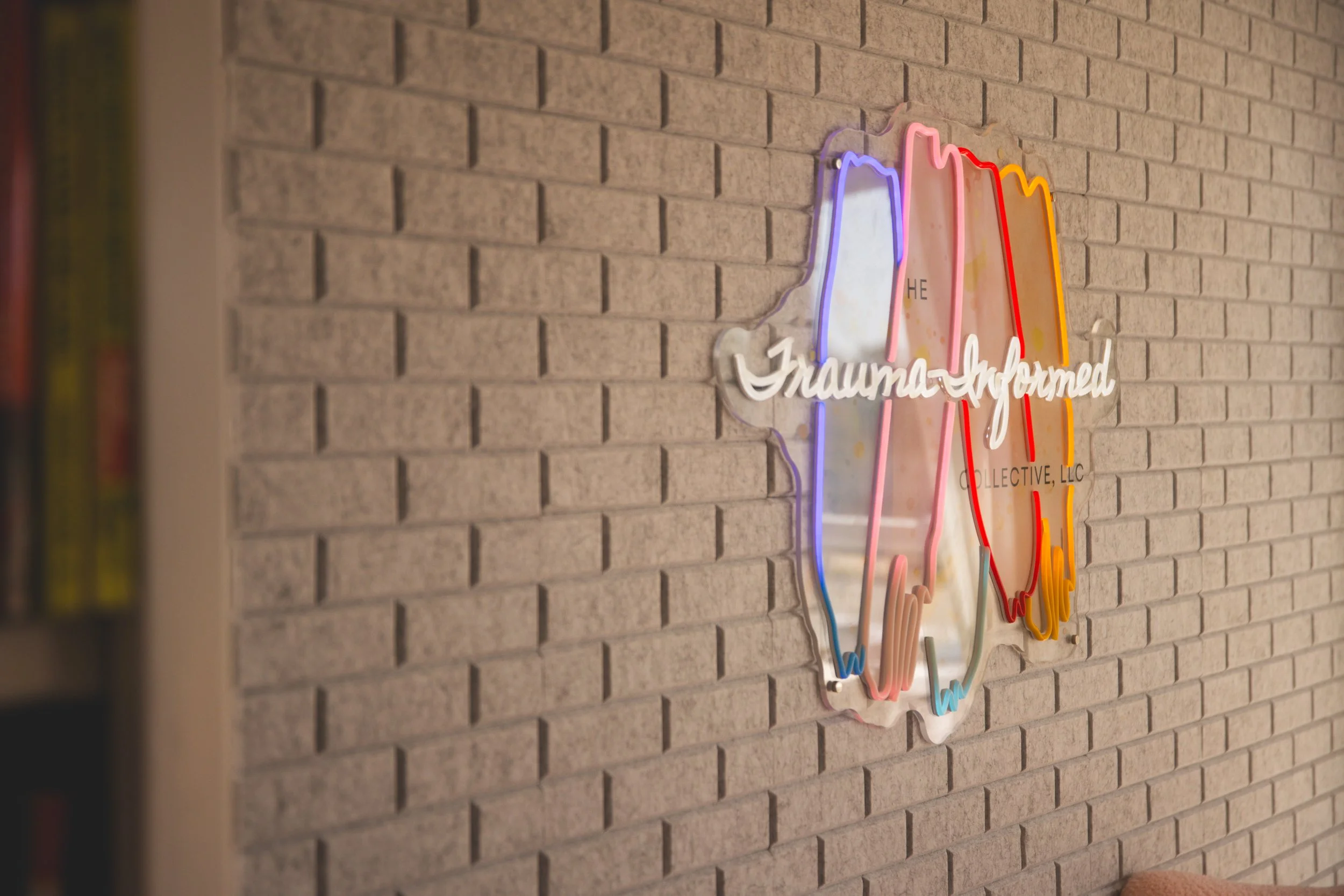 Colorful neon sign on a brick wall, with the phrase 'The Trauma-Informed Collective, LLC' and a stylized outline of a brain.