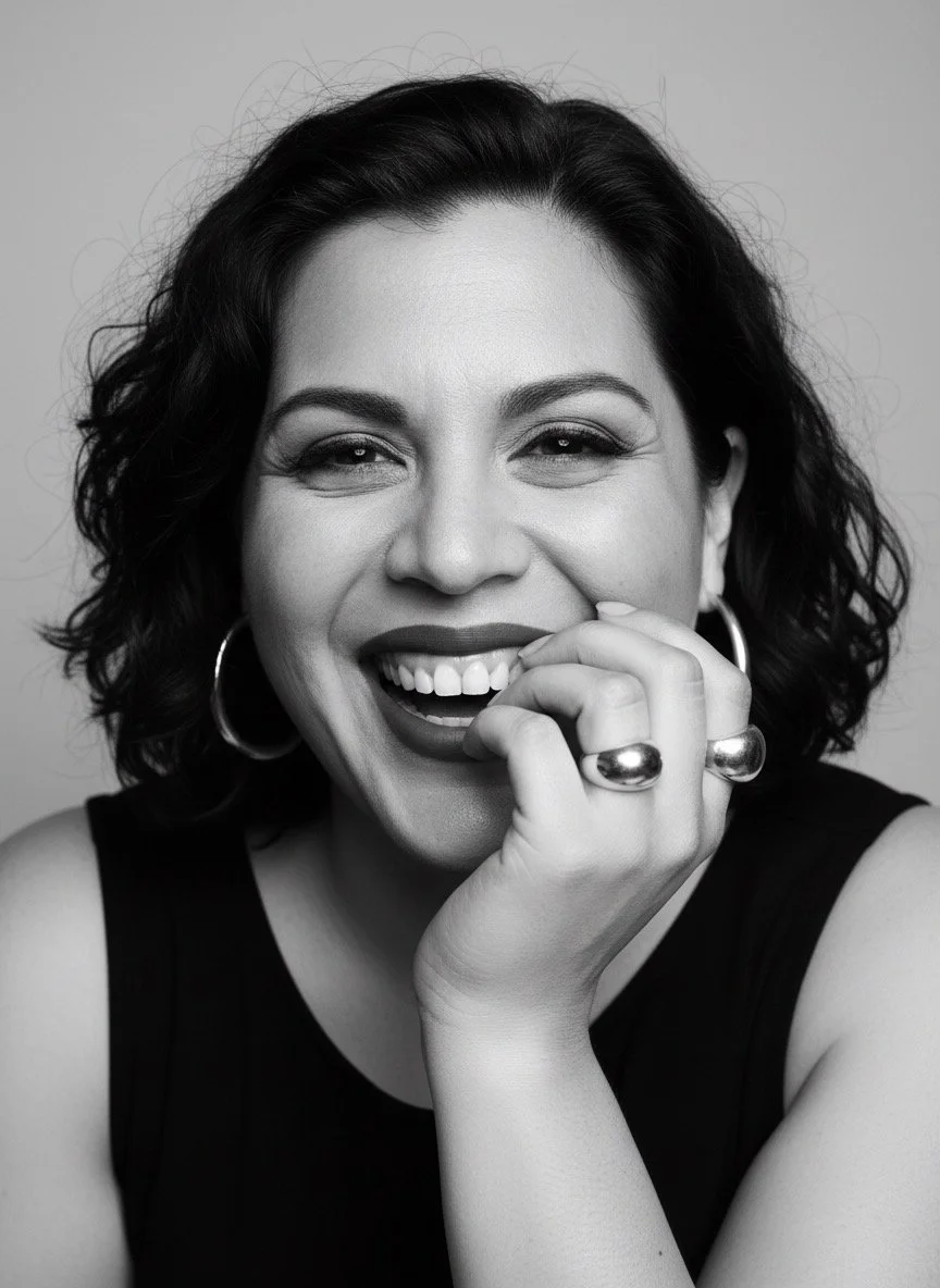 Black and white portrait of a woman smiling warmly with her hand near her chin, wearing large hoop earrings and bold rings.
