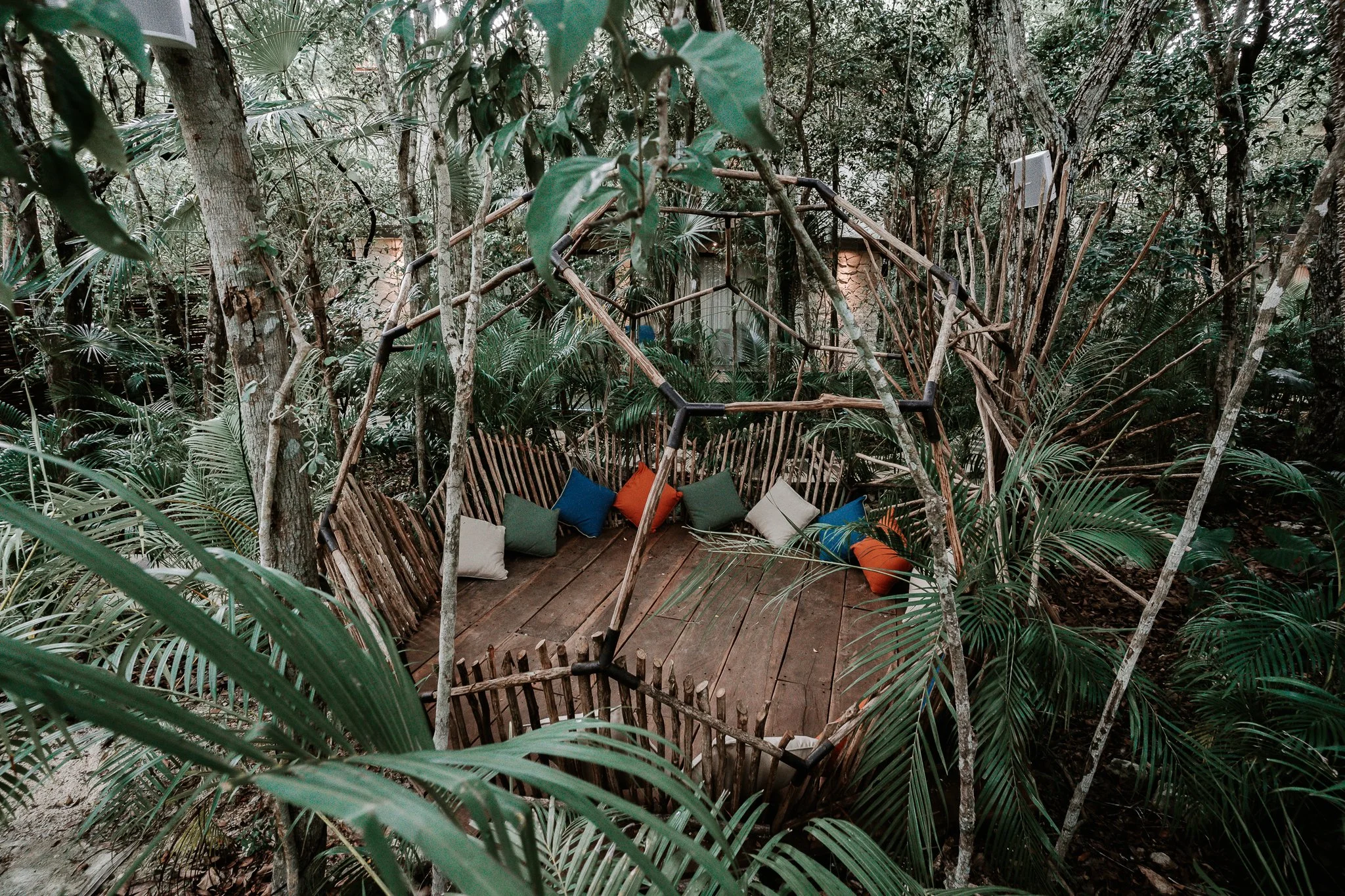 A wooden deck in a lush, tropical forest with a rustic swing made of branches and ropes, decorated with colorful cushions, surrounded by dense green foliage.
