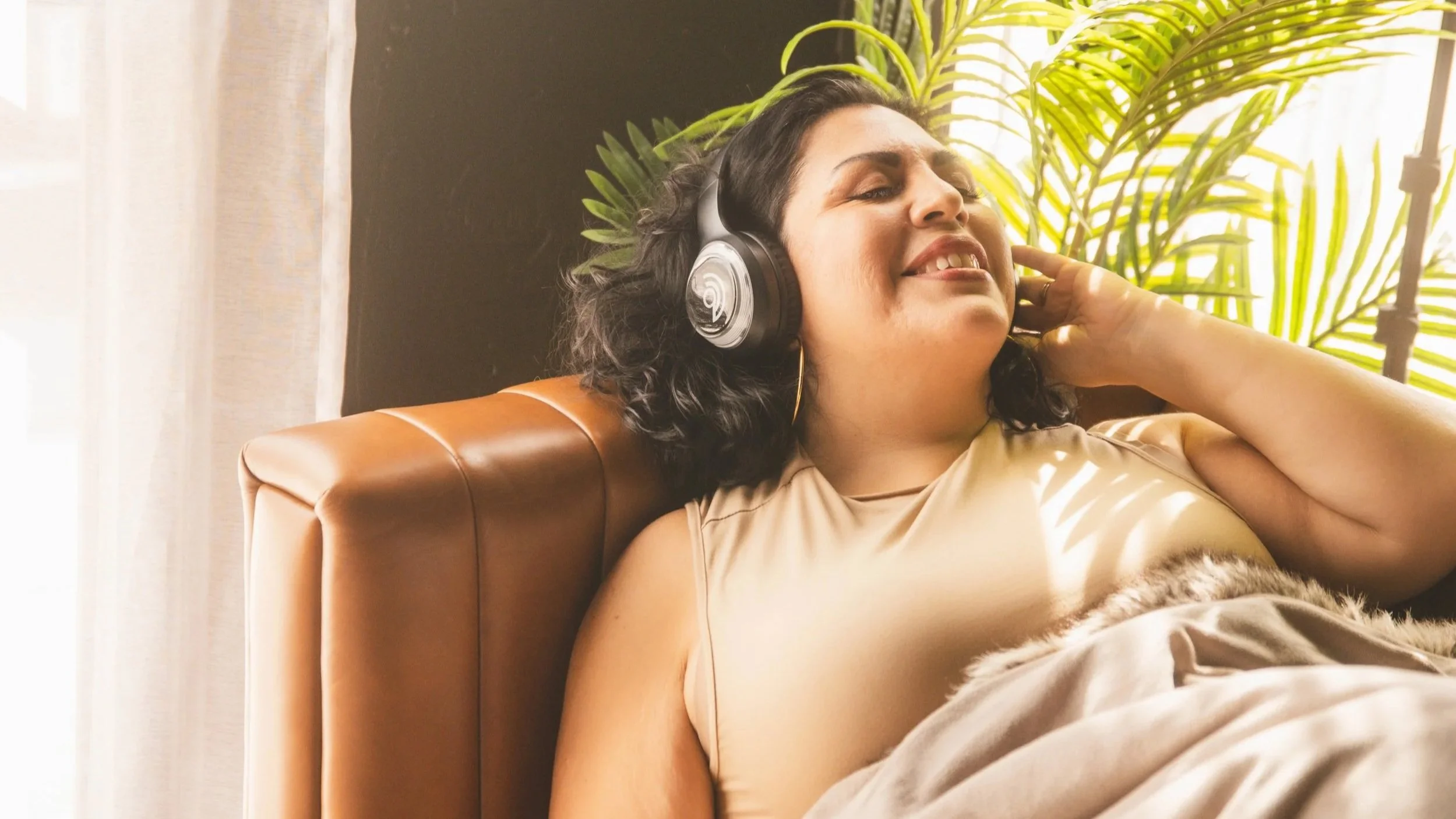 A woman with dark curly hair smiling and relaxing with her eyes closed, wearing headphones indoors, with a plant and natural sunlight in the background.
