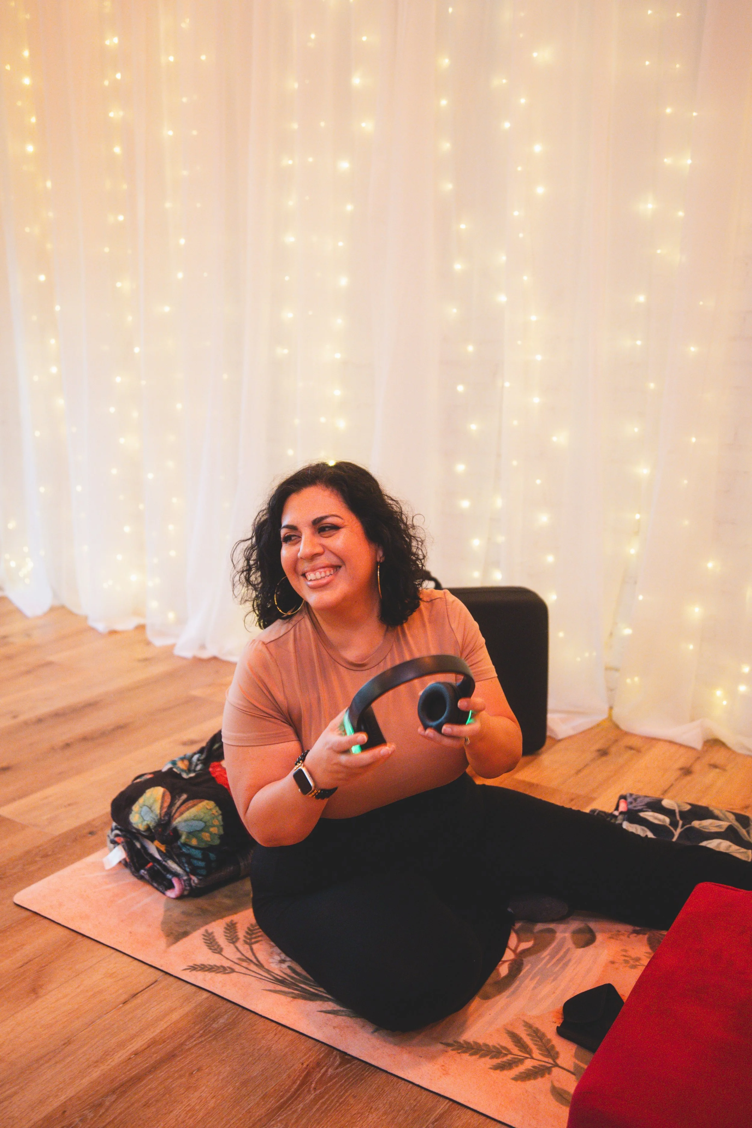 A woman with curly dark hair, smiling, sitting cross-legged on a yoga mat indoors. She is holding headphones and wearing a smartwatch. Behind her is a white curtain with string lights, and on the floor next to her is a colorful bag.