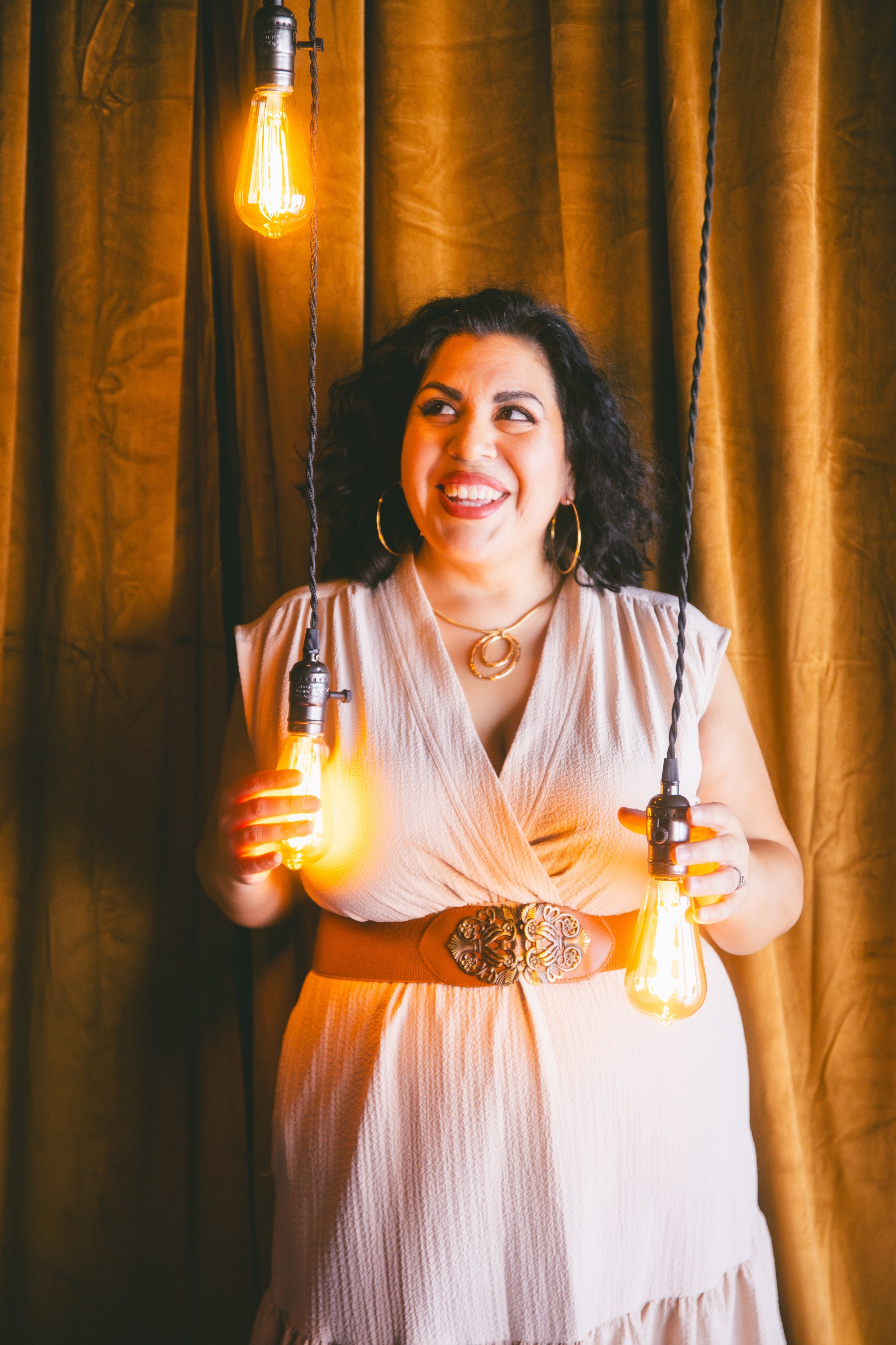 A woman with curly dark hair, wearing gold hoop earrings, a necklace, and a beige dress with a wide belt, holding two illuminated light bulbs with black cords, standing in front of gold curtains.