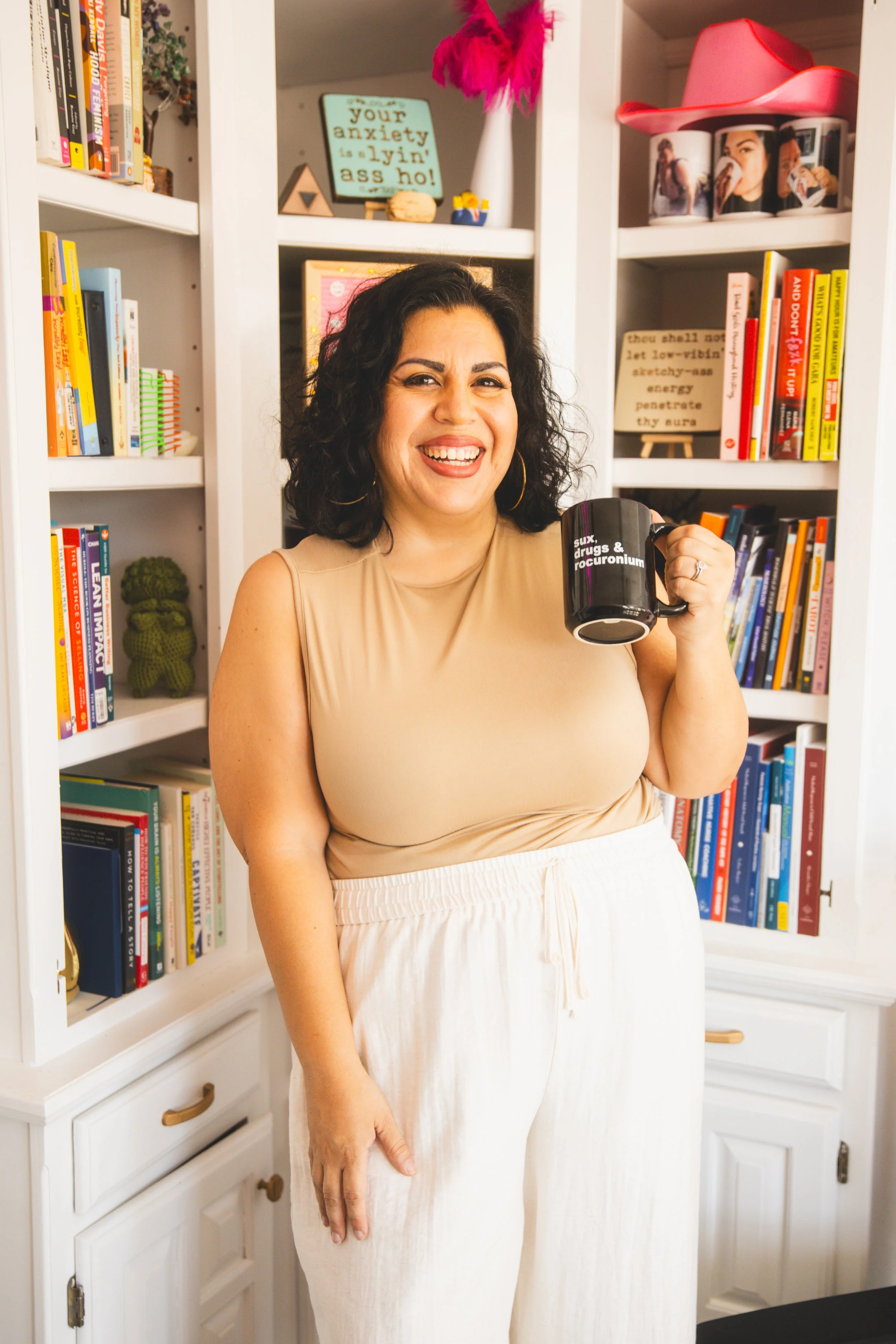 Women smiling, holding a black coffee mug with the text 'sux, drugs & roocuronium', standing in front of white bookshelves filled with books, photos, and decorative items.