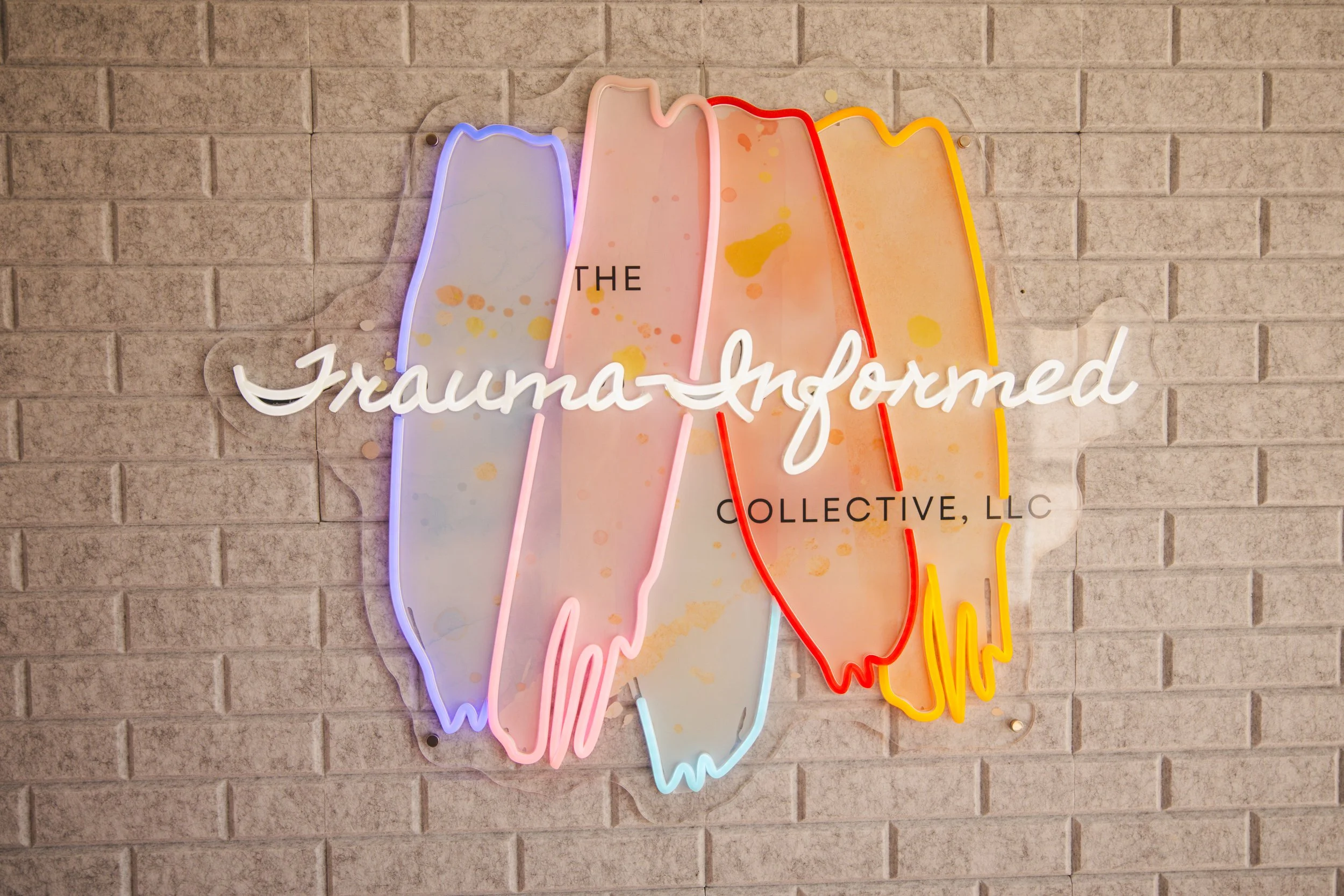 A neon sign on a brick wall spelling out "The Traumatized Collective, LLC" with colorful abstract shapes resembling stylized driftwood or paint splatters.
