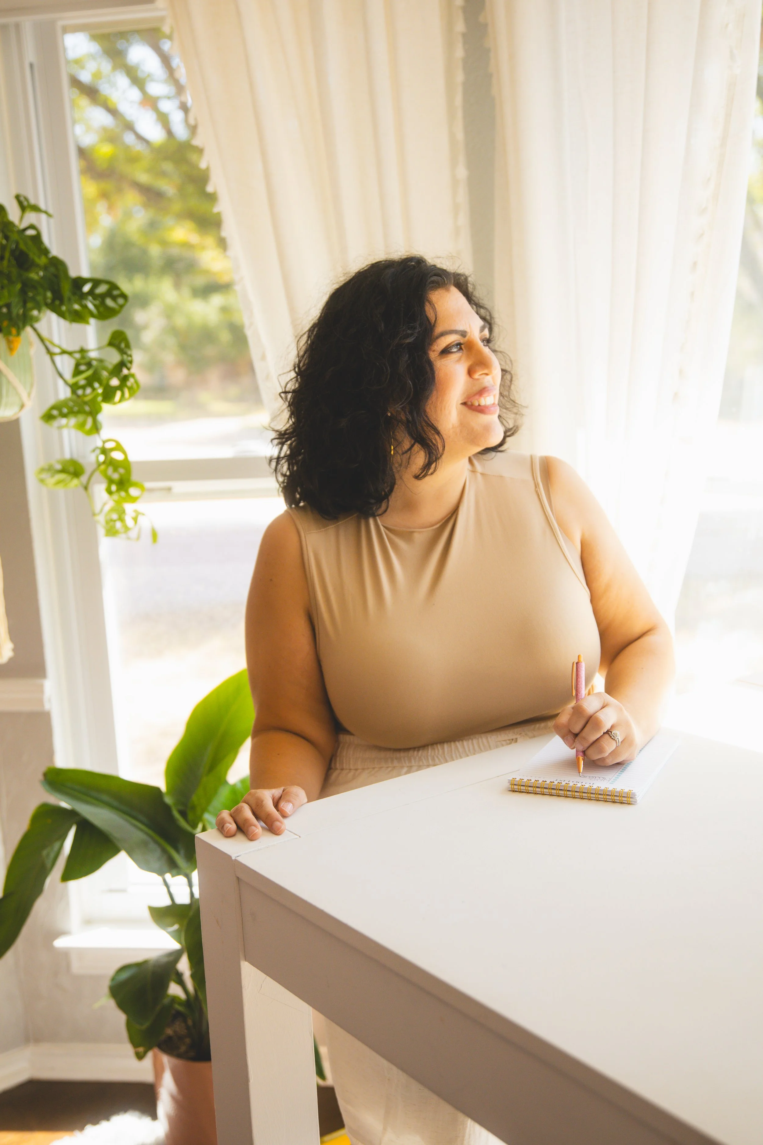 A woman with dark curly hair seated at a white table, smiling, holding a pen over a notepad, with large windows, white curtains, and green plants in the background.