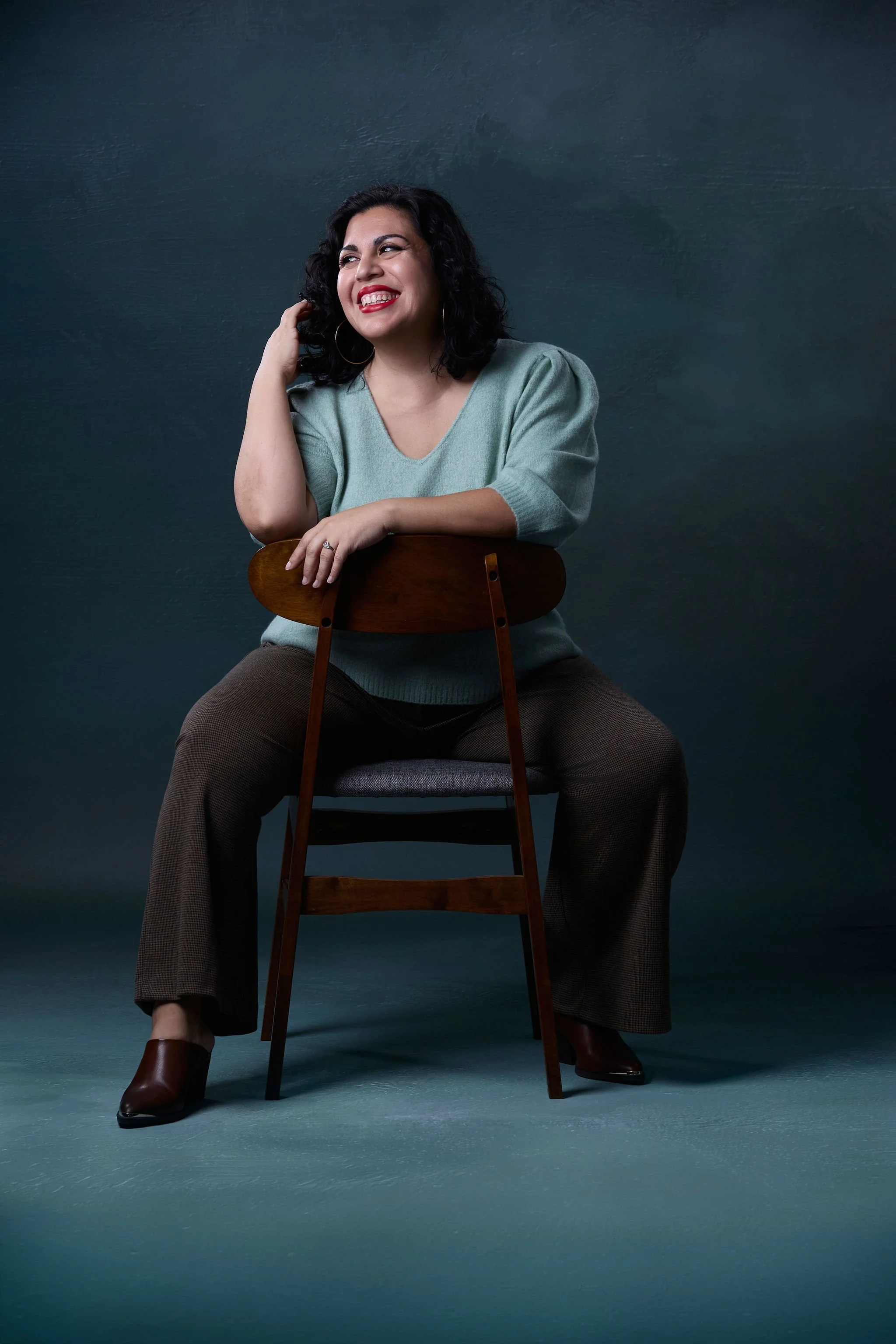 A woman with dark curly hair, wearing a mint green top, brown plaid pants, and brown boots, sitting on a wooden chair against a dark teal background, smiling and looking to her left.