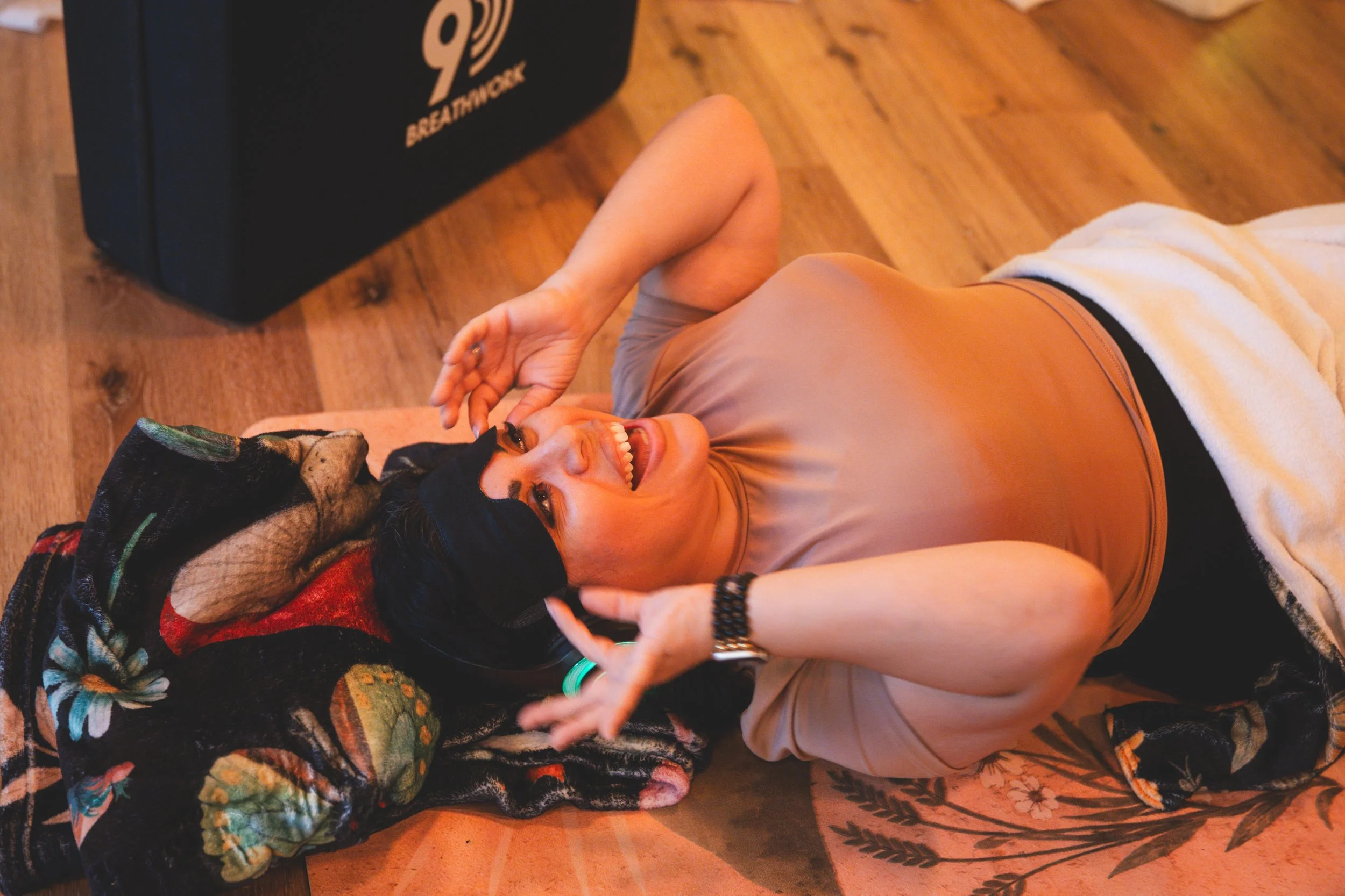 A woman lying on the floor, smiling and holding her nose, with an older person resting on her chest, both wearing casual clothes, and the floor made of wood. The woman has dark hair and a watch on her wrist, and the older person is wrapped in a colorful blanket.
