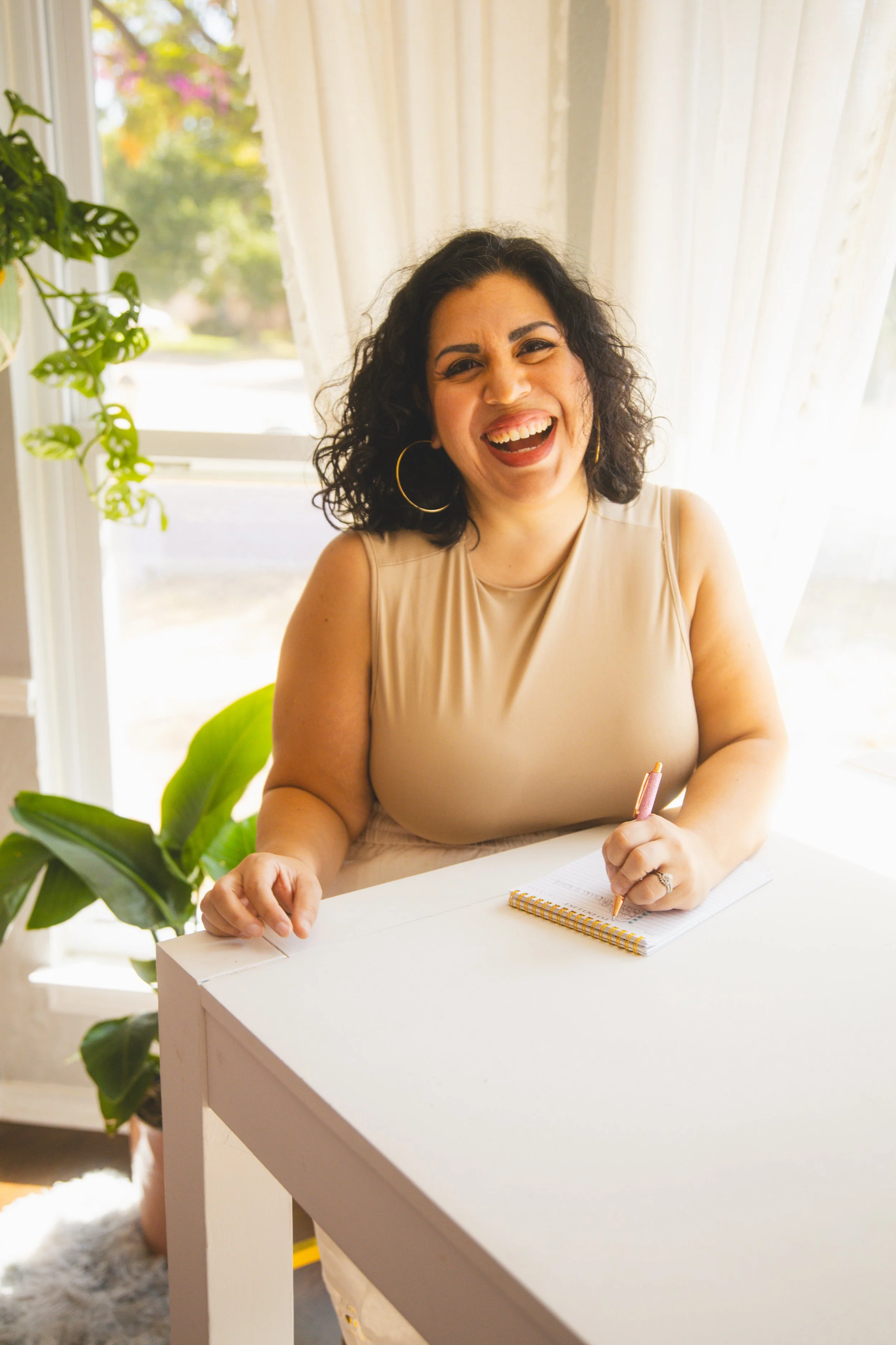 A woman with dark, curly hair sitting at a white table, smiling and holding a pen over a notebook, with houseplants and curtains in the background.