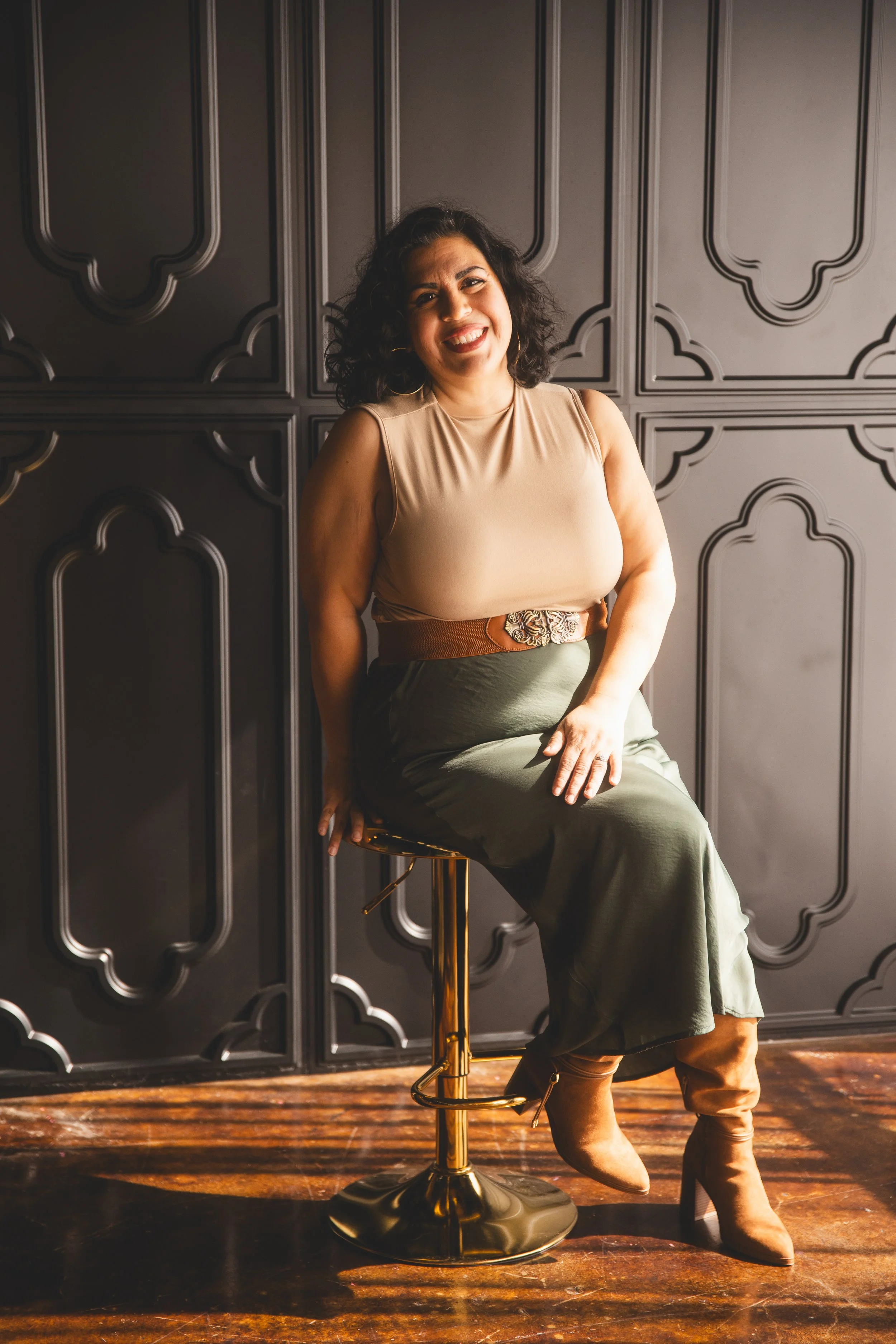 A woman with curly dark hair sitting on a bar stool, smiling at the camera, in front of a dark decorative wall, wearing a beige sleeveless top and a green skirt.