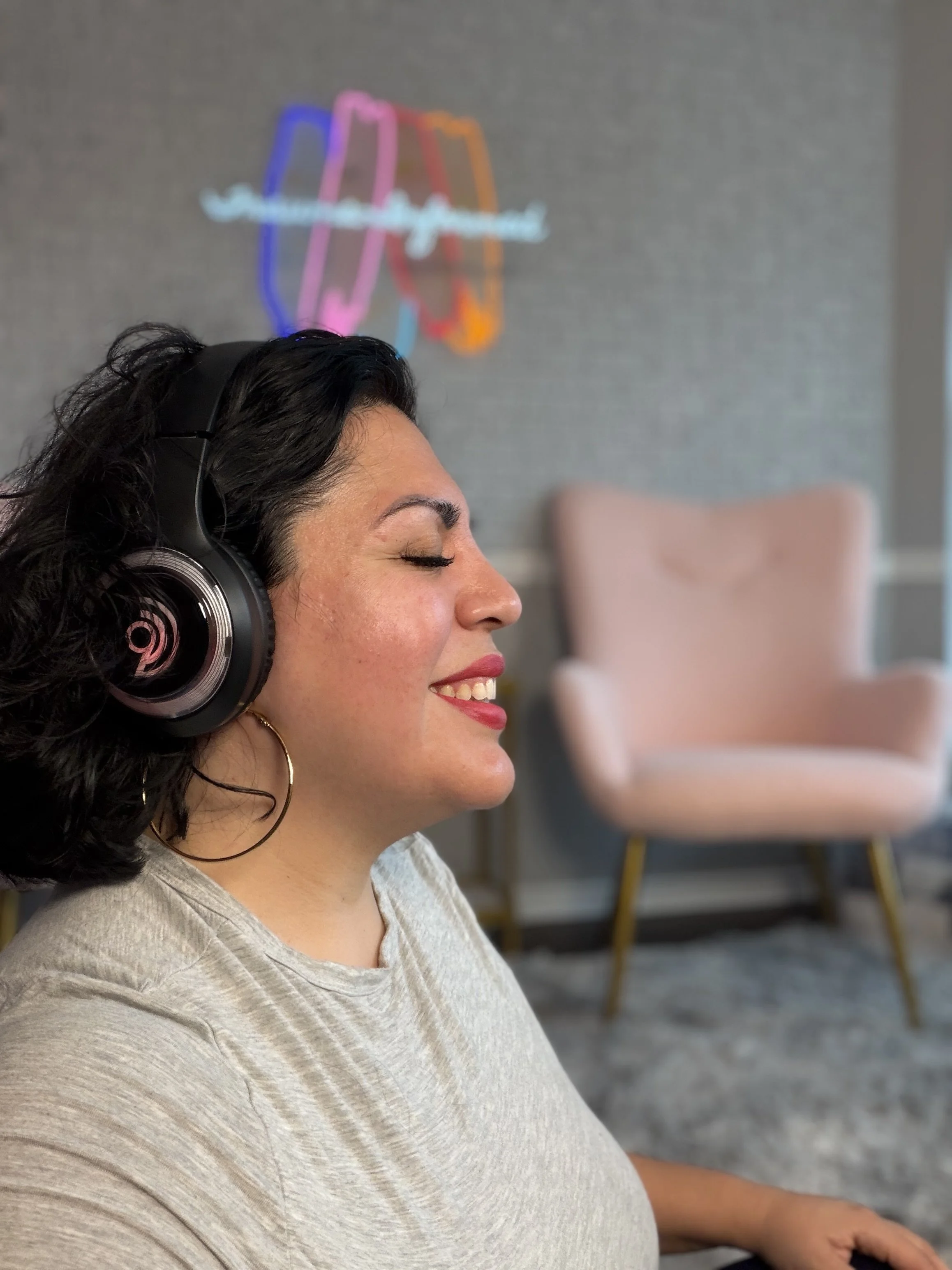 A woman with black curly hair and hoop earrings wearing headphones, smiling with eyes closed, in a room with a pink armchair and a neon sign on the wall in the background.