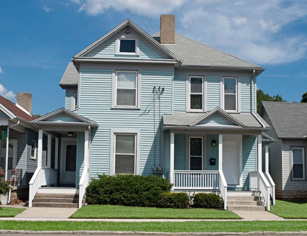 A light blue, multi-story house with a gabled roof, front porch, front steps, and surrounding green lawn