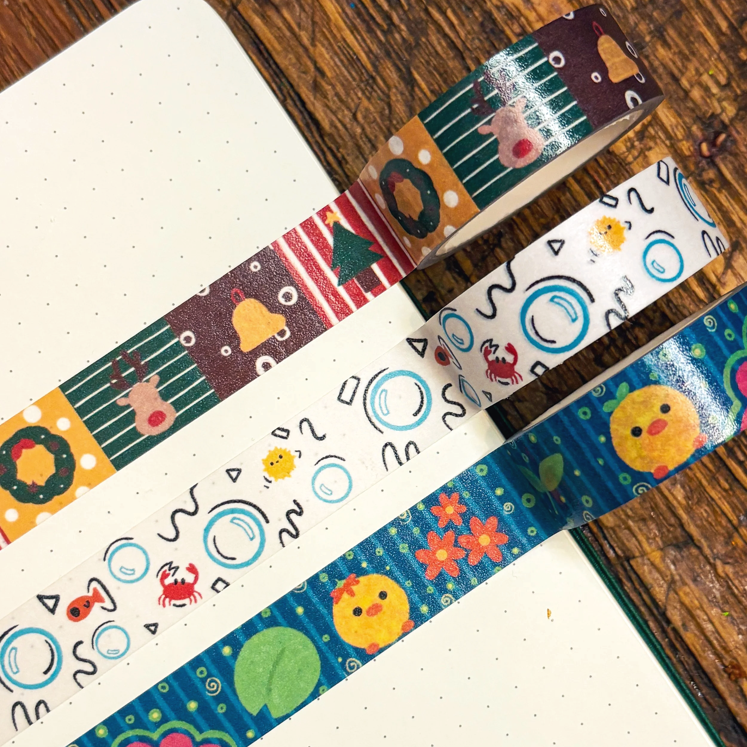 Decorative washi tape rolls with Christmas and holiday-themed patterns, including reindeer, wreaths, snowflakes, a chick, and floral motifs, placed on dotted notebooks on a wooden surface.