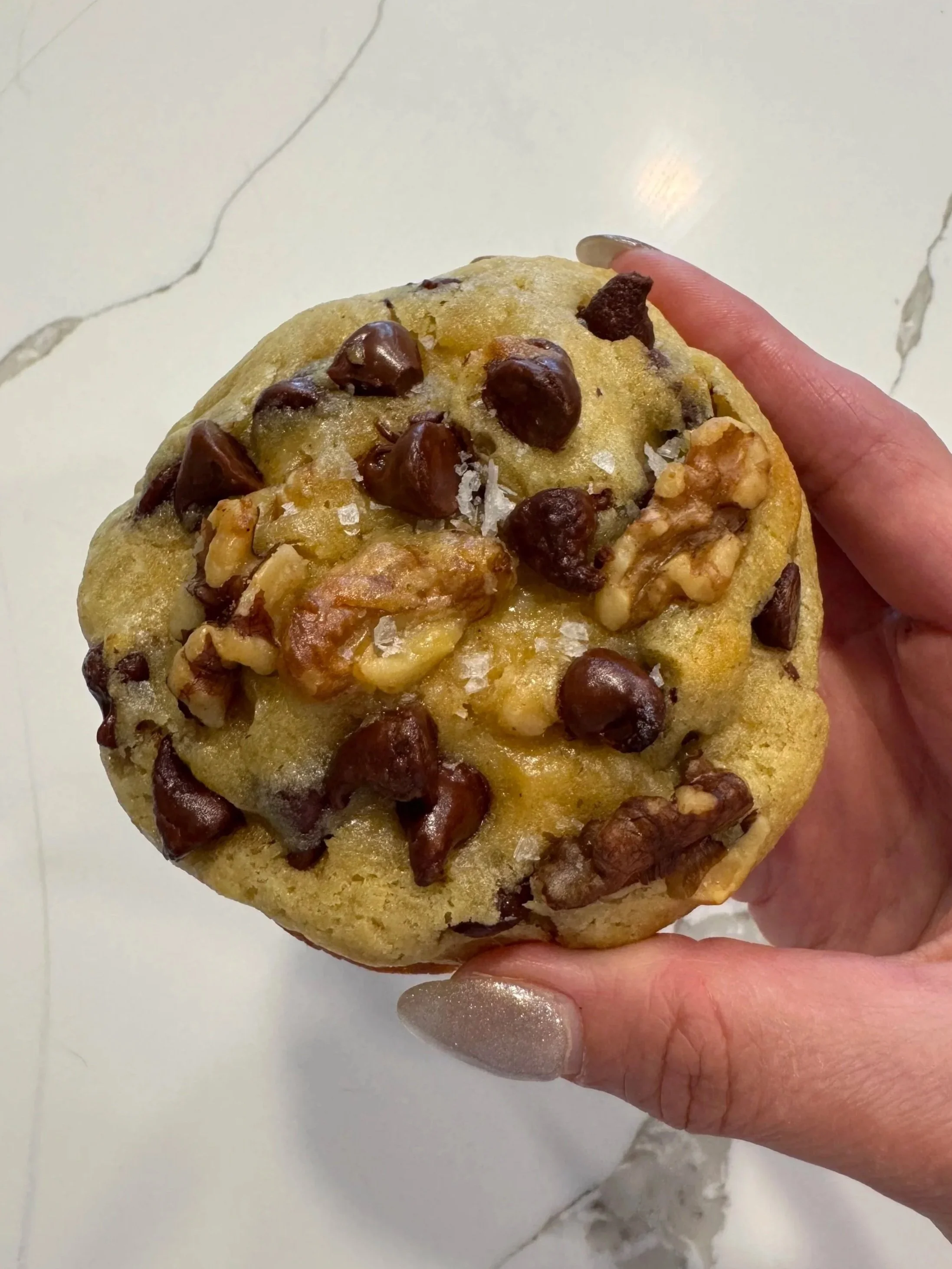 Hand holding a large chocolate chip cookie with walnuts, on a white marble surface.