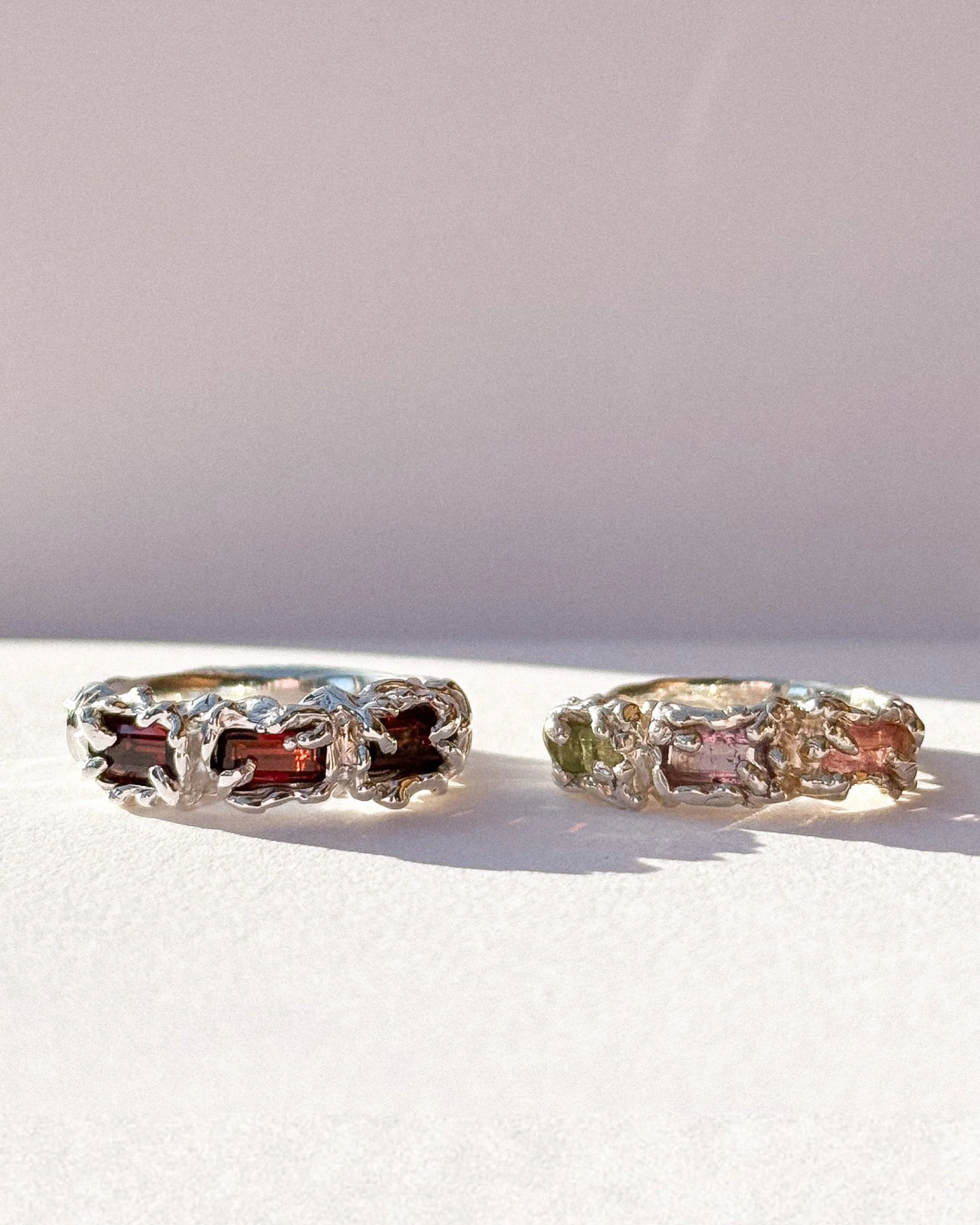 Two silver rings with rectangular red, pink, and green gemstones, set in intricate metalwork, placed on a white surface with a neutral background.