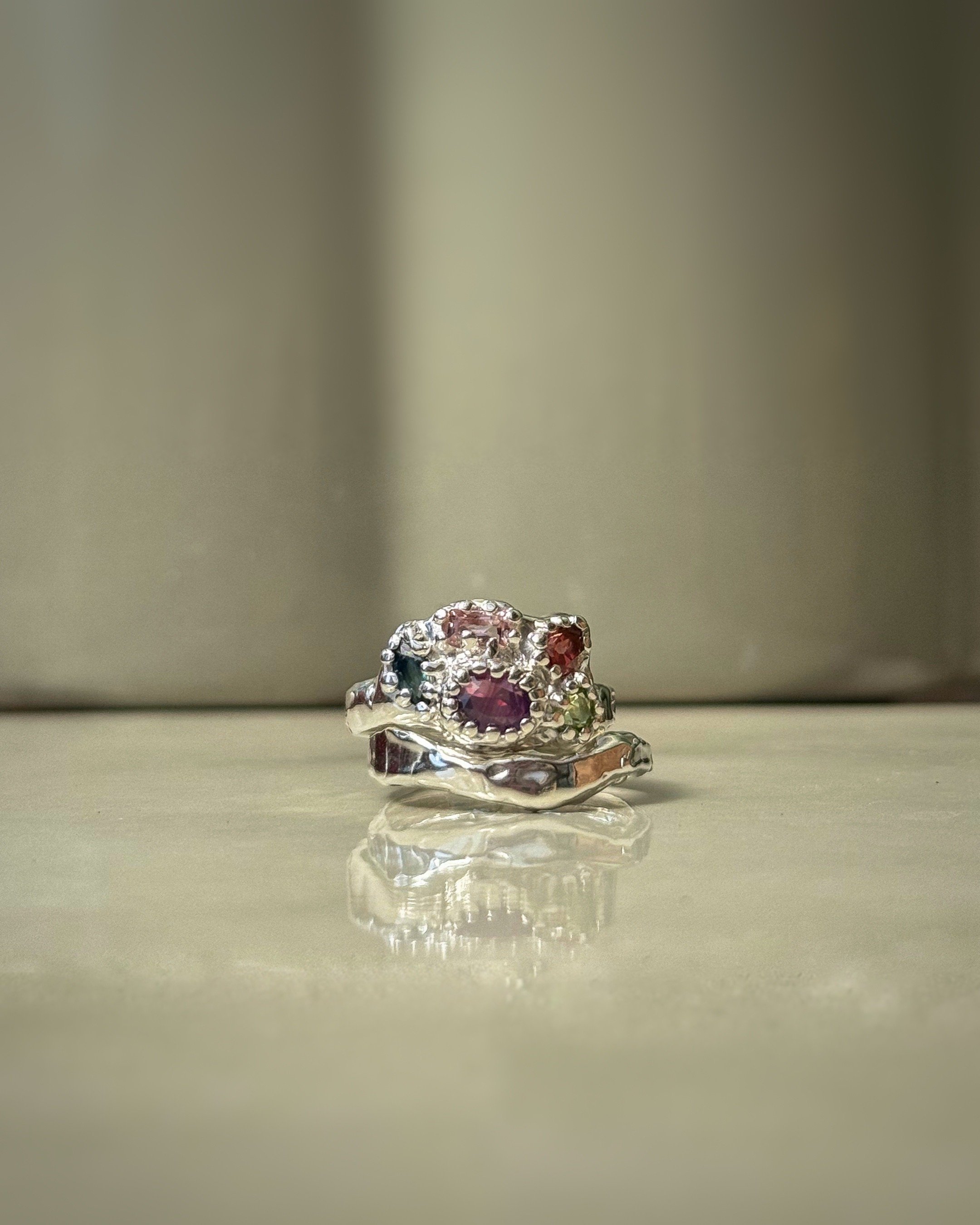 Close-up of a silver ring with multiple small, colorful gemstones, resting on a reflective surface.
