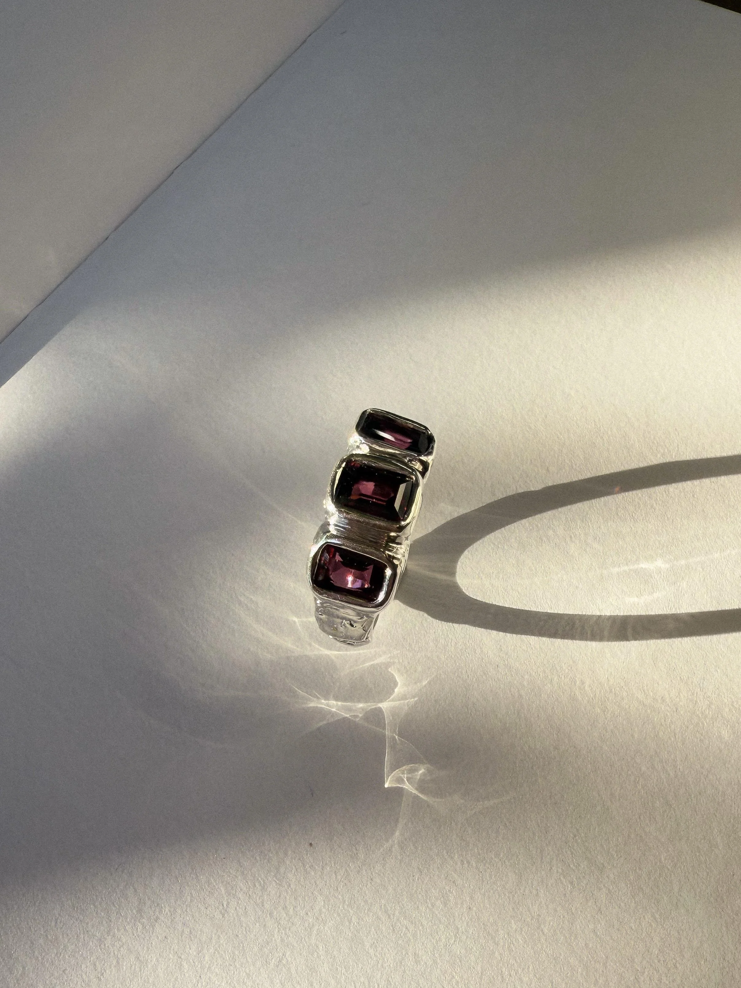 Silver ring with four square purple gemstones on a white surface, casting a circular shadow.