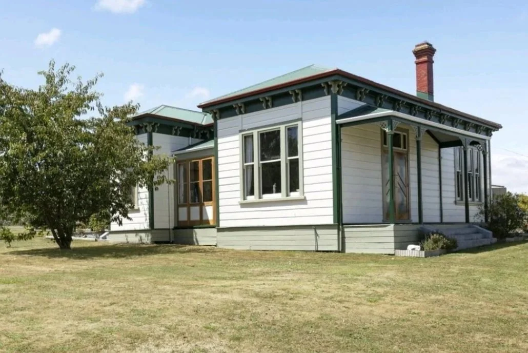 A two-story house with white siding, green trim, and a brick chimney, surrounded by a lawn and a tree.