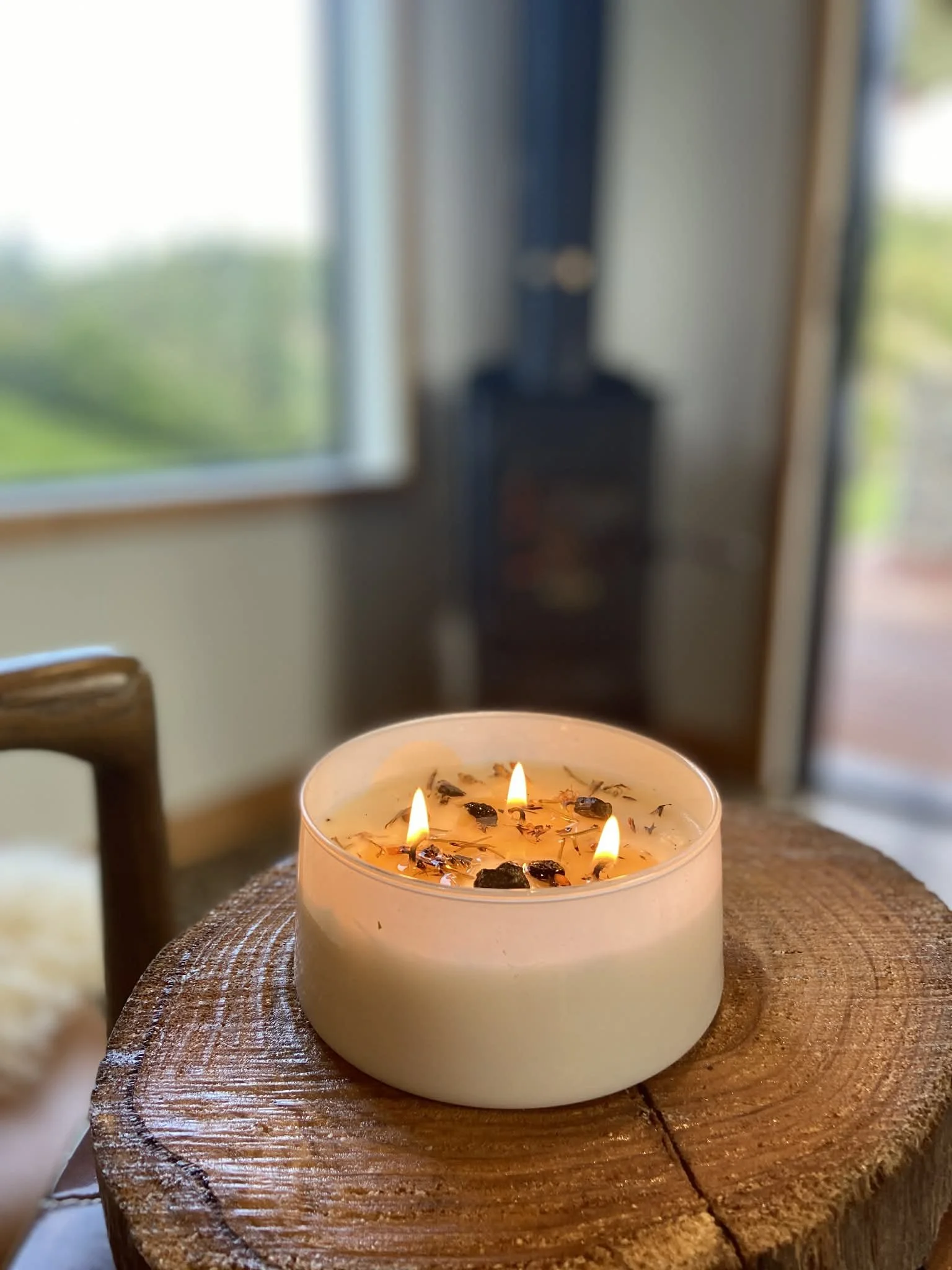 A white candle with three flames on a wooden tabletop, indoors near a window with a blurry view outside.