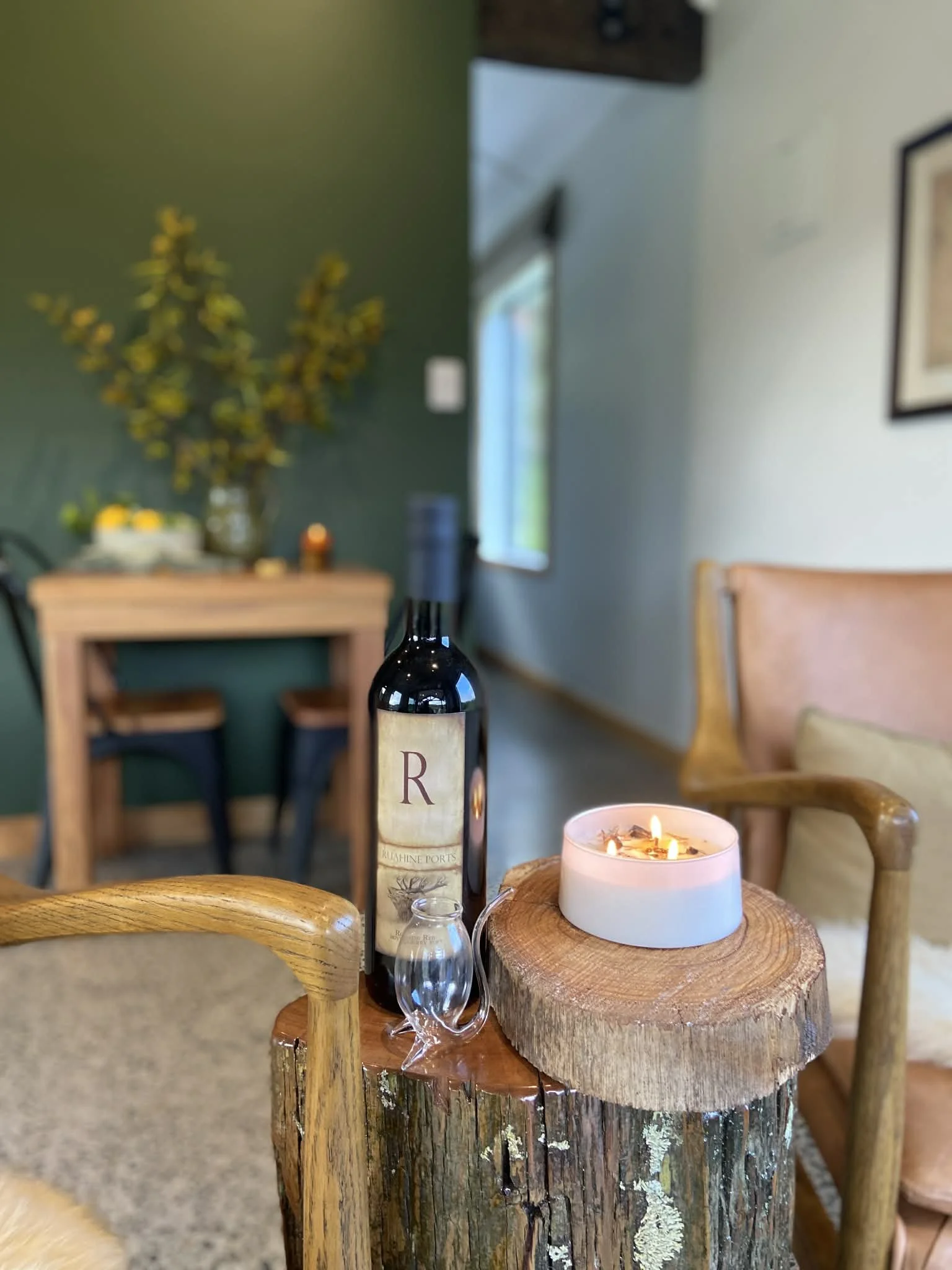 A cozy indoor scene with a wooden table holding a lit candle, a bottle of wine, and a wine glass. There are chairs around a rustic wooden stump table. In the background, there is a green accent wall, a shelf with plants and decor, and a window letting in natural light.
