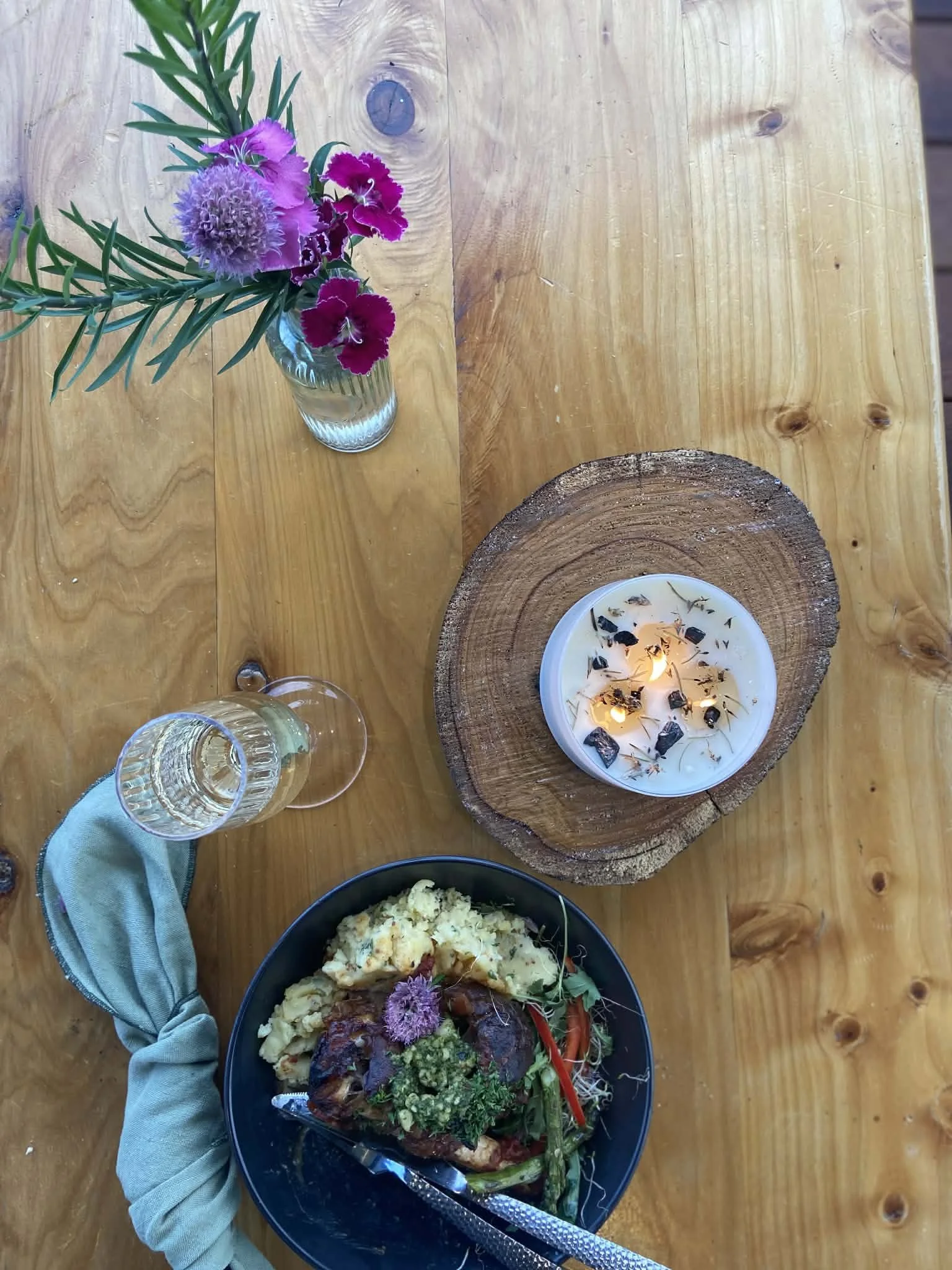 A wooden table with a vase of purple, pink, and green flowers, a black bowl of food with mashed potatoes, vegetables, and meat, a lit candle on a wooden base, and a glass of white wine