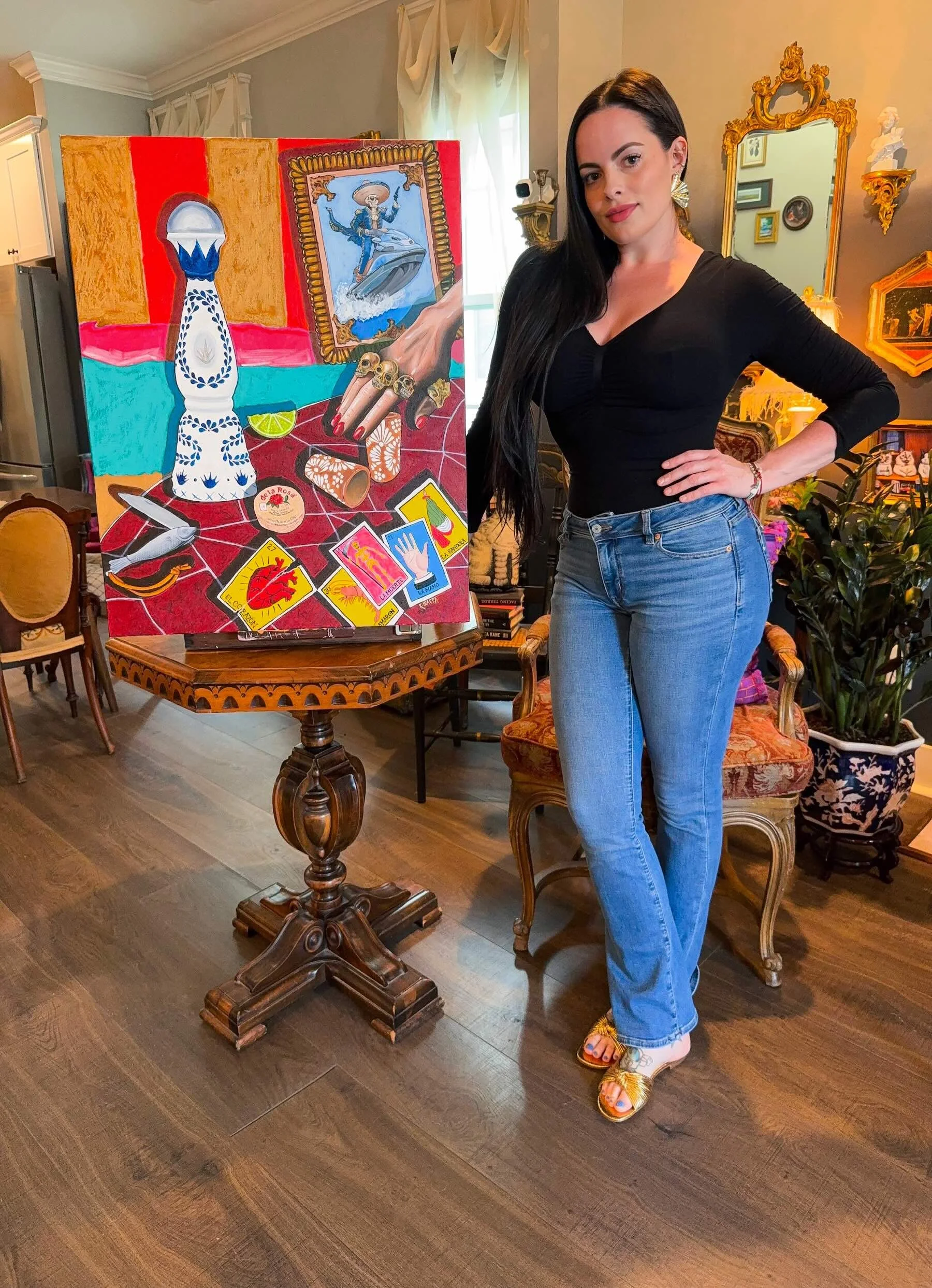 A woman with long black hair, wearing a black top, blue jeans, and colorful sandals, stands next to an easel with a brightly colored painting in a decorated living room with vintage furniture and wall decorations.