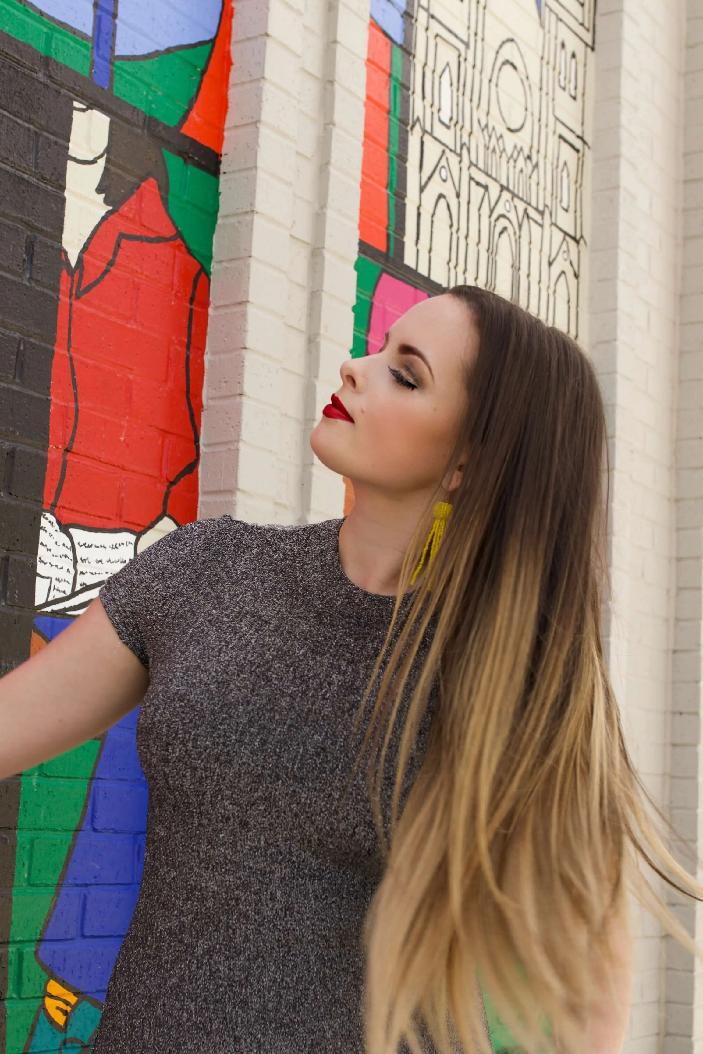 A woman with long blonde hair and red lipstick leaning against a colorful mural on a brick wall, with a relaxed expression.