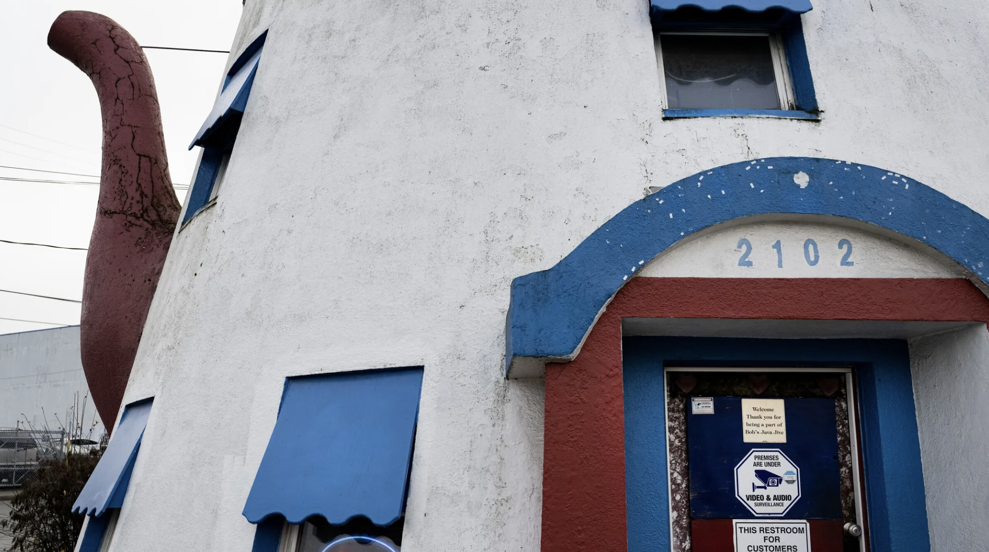 A white building shaped like a teapot with blue and red accents designed to look like a whimsical cartoon house, featuring a large red chimney, blue window awnings, and a colorful curved facade.