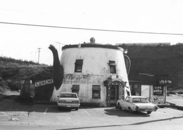 A building designed to look like a large coffee pot, with parked cars in front and a sign for lunch and live music in the background.