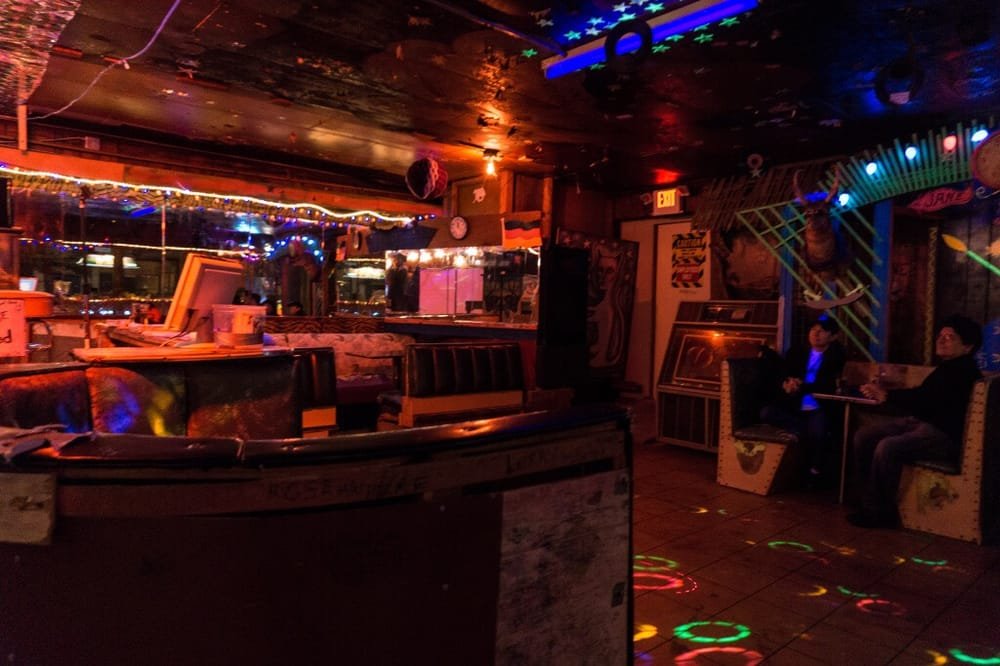 Interior of a dimly lit bar with colorful decorations, string lights, and neon lights. Two people are sitting on the right side, conversing, with arcade machines visible in the background.