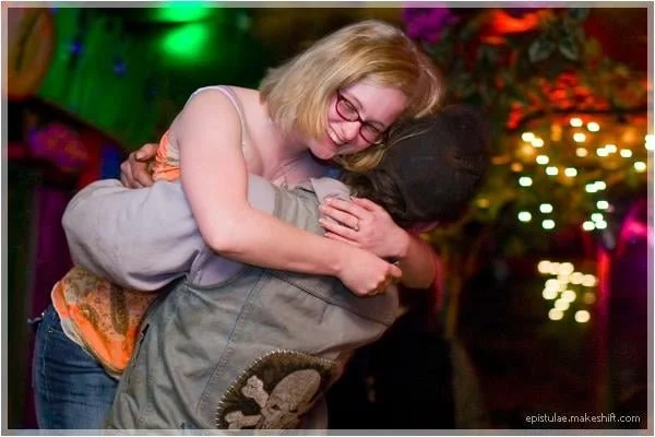Two women hugging, one smiling with glasses and blonde hair, the other with dark hair facing away, in a colorful, festive setting with lights.