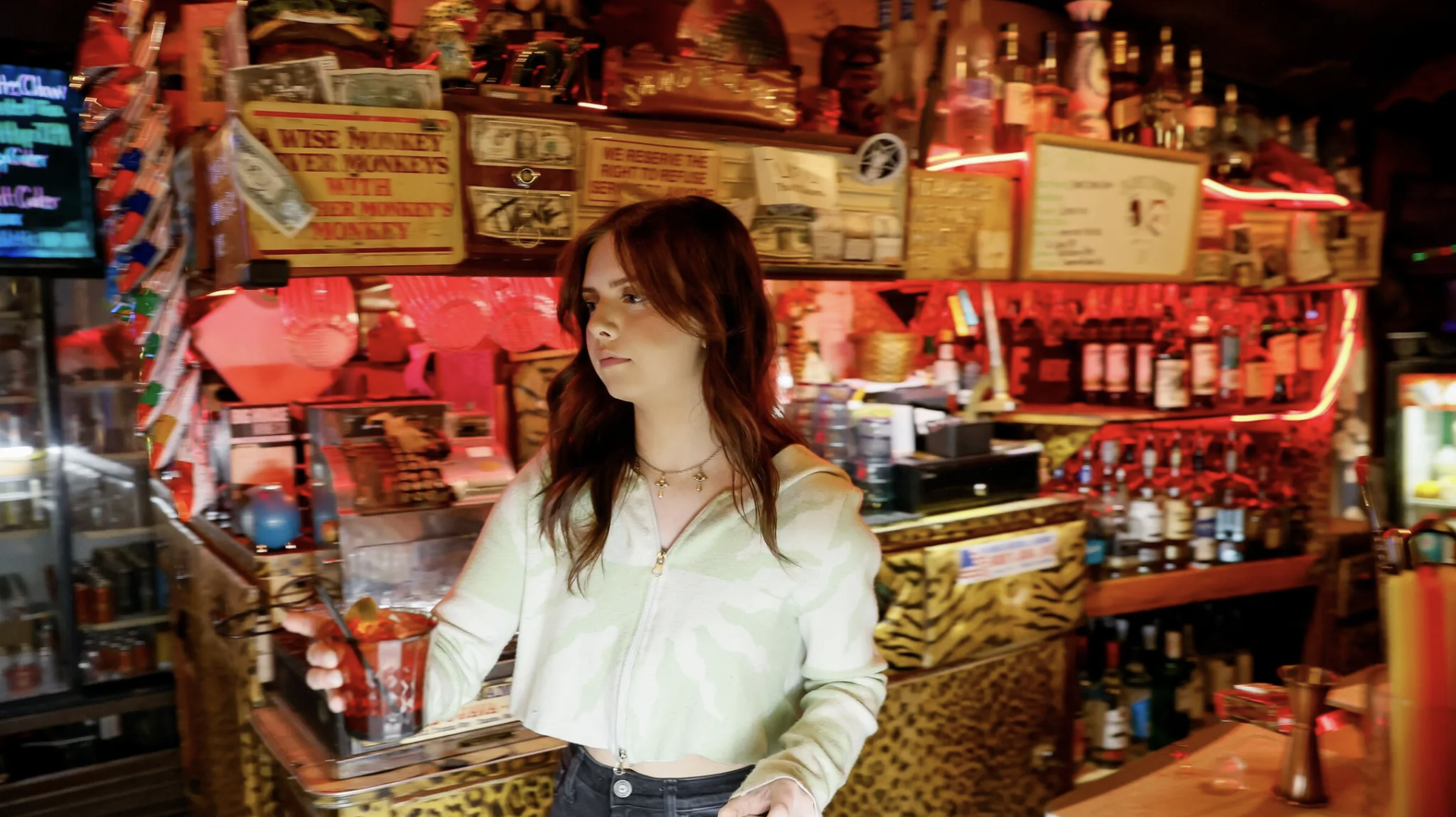 A woman with reddish-brown hair wearing a light-colored zip-up jacket and necklace, holding a drink with a straw in a bar with red and warm lighting, surrounded by bottles and bar decorations.