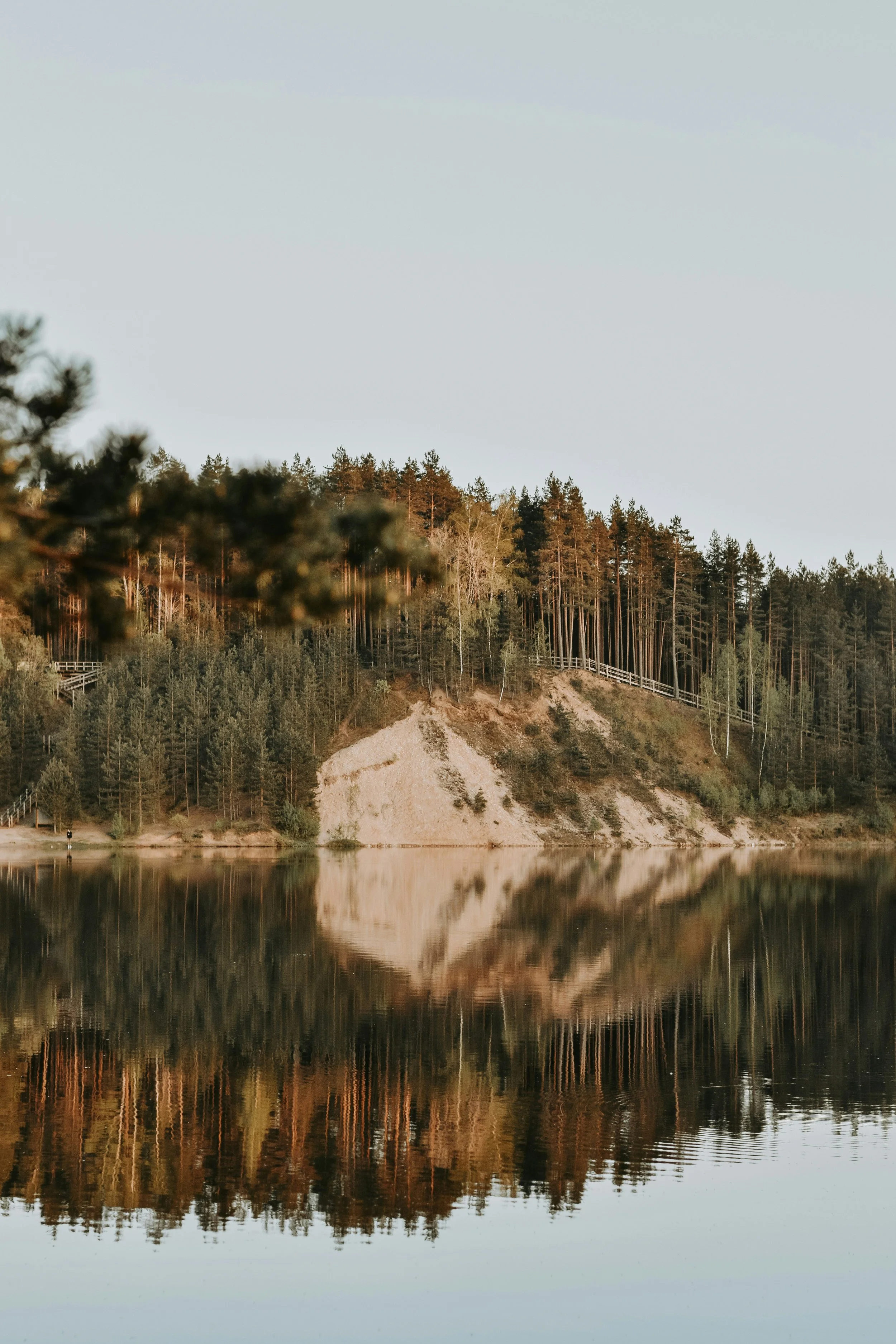 A calm lake reflecting a hillside with trees and sandy patches, under a cloudy sky.