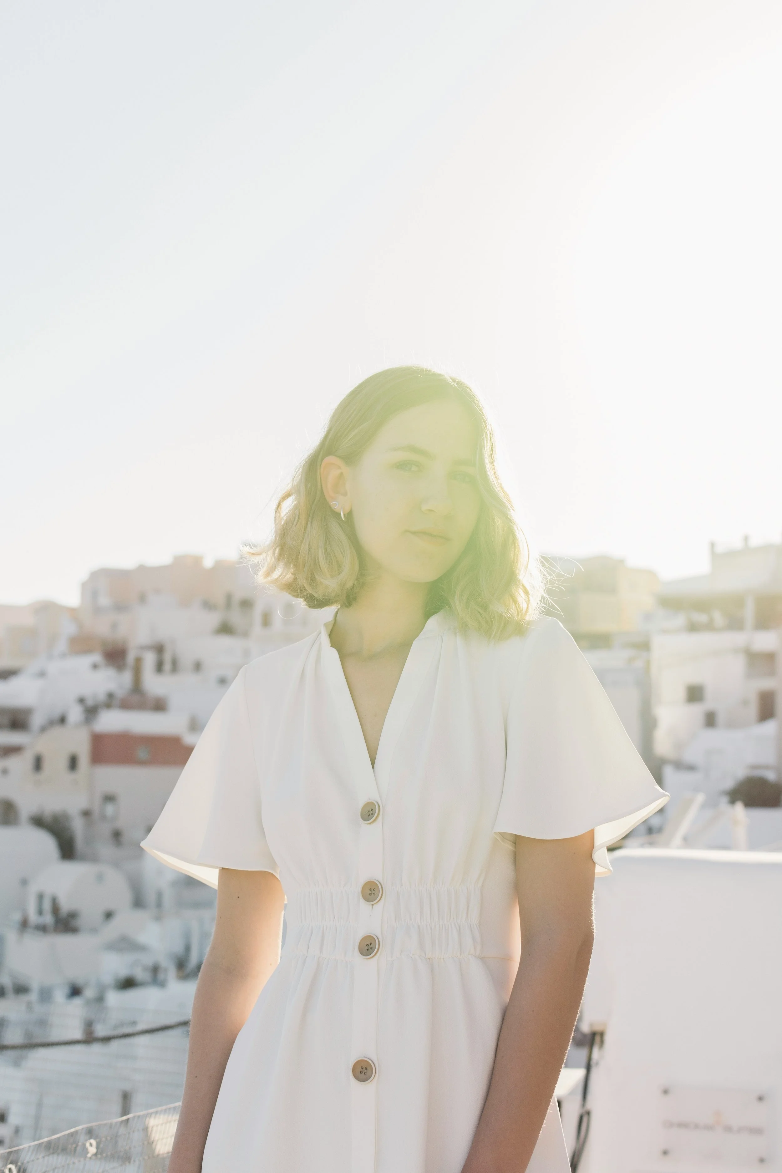 A young woman in a white dress stands outdoors in front of white buildings, illuminated by sunlight.