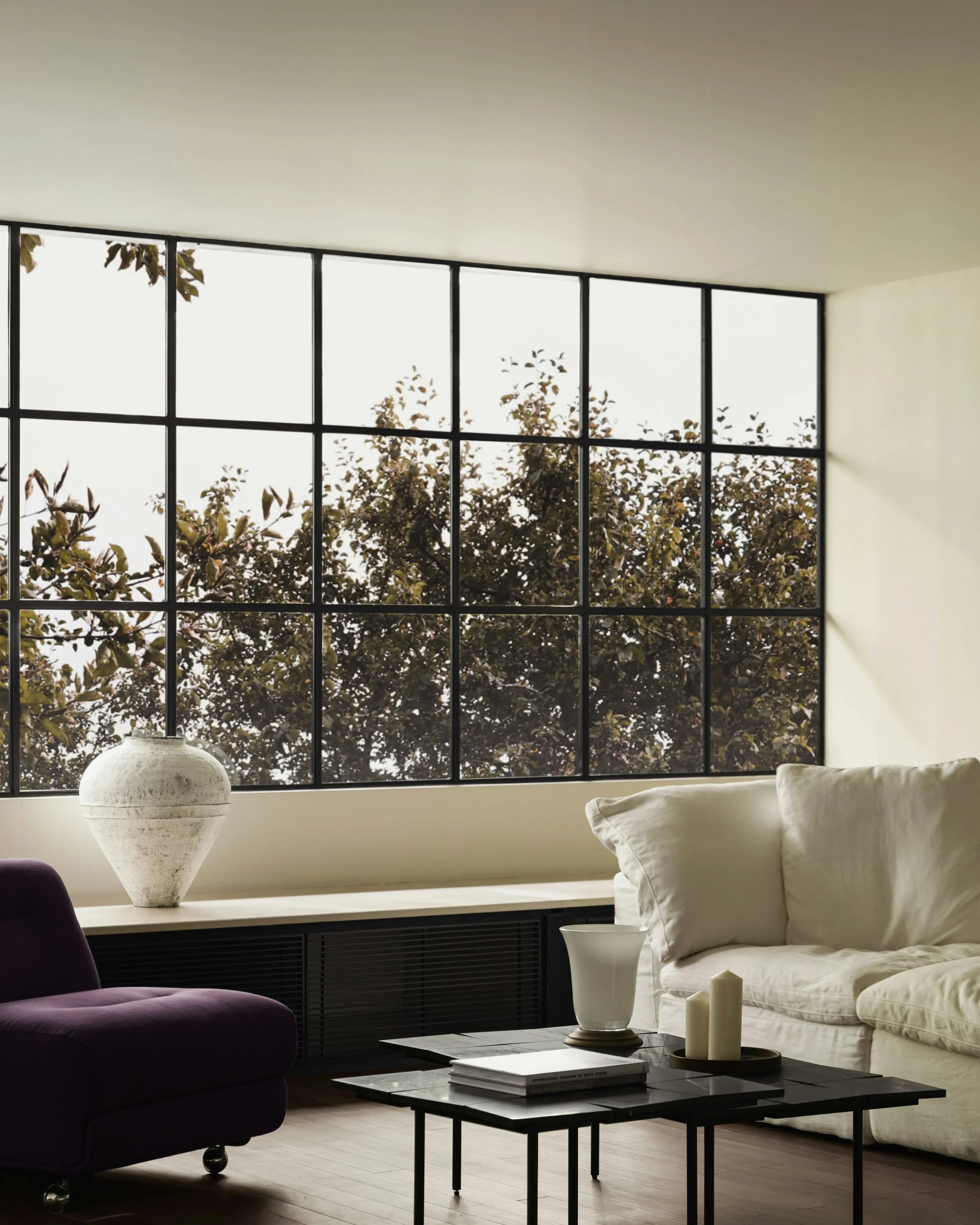 Living room with a large window, white couch, dark purple chair, coffee table with candles and books, large vase with greenery, and a window view of trees outside.
