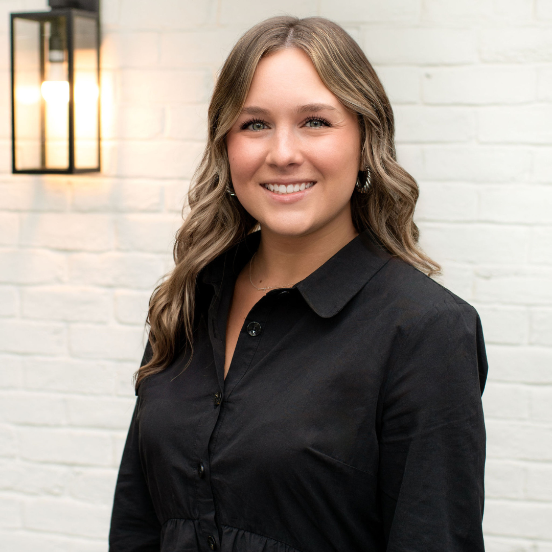 A woman with wavy light brown hair smiling, wearing a black button-up shirt, standing in front of a white brick wall with a lit wall sconce.