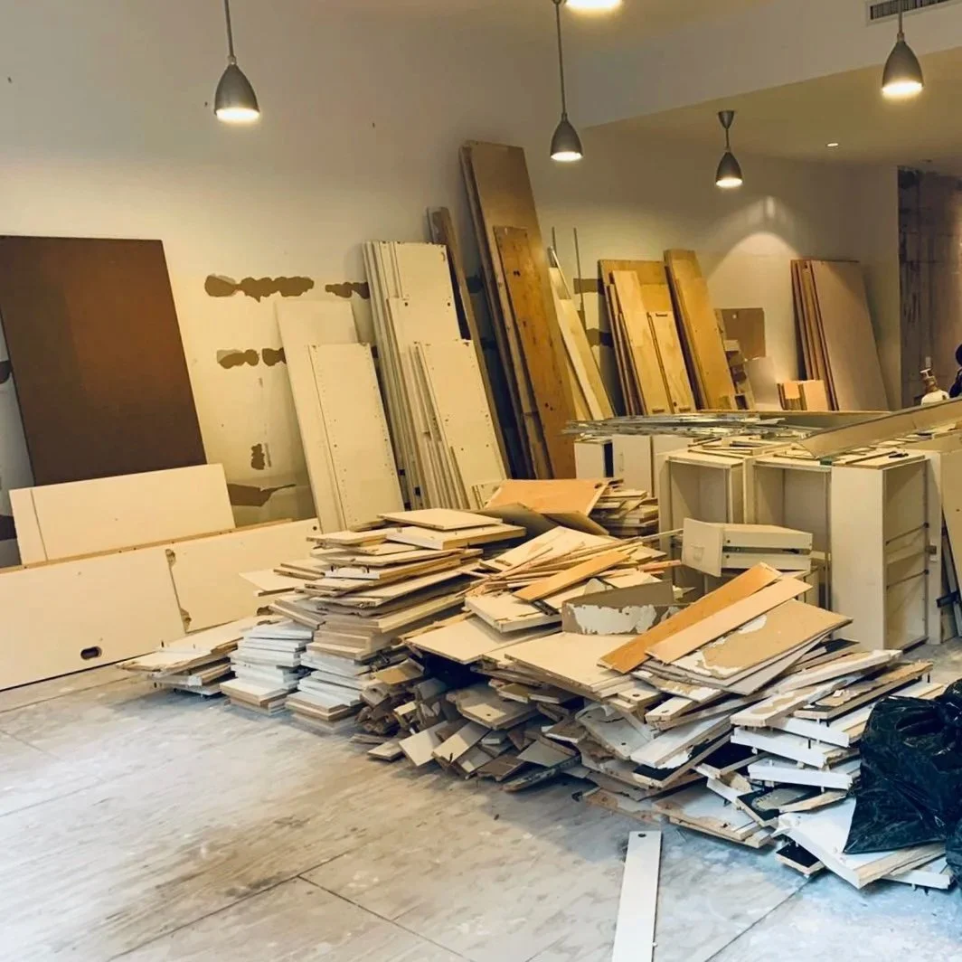 Piles of cardboard and wooden boards stacked in an unfinished or under-construction room with white walls and hanging lights.