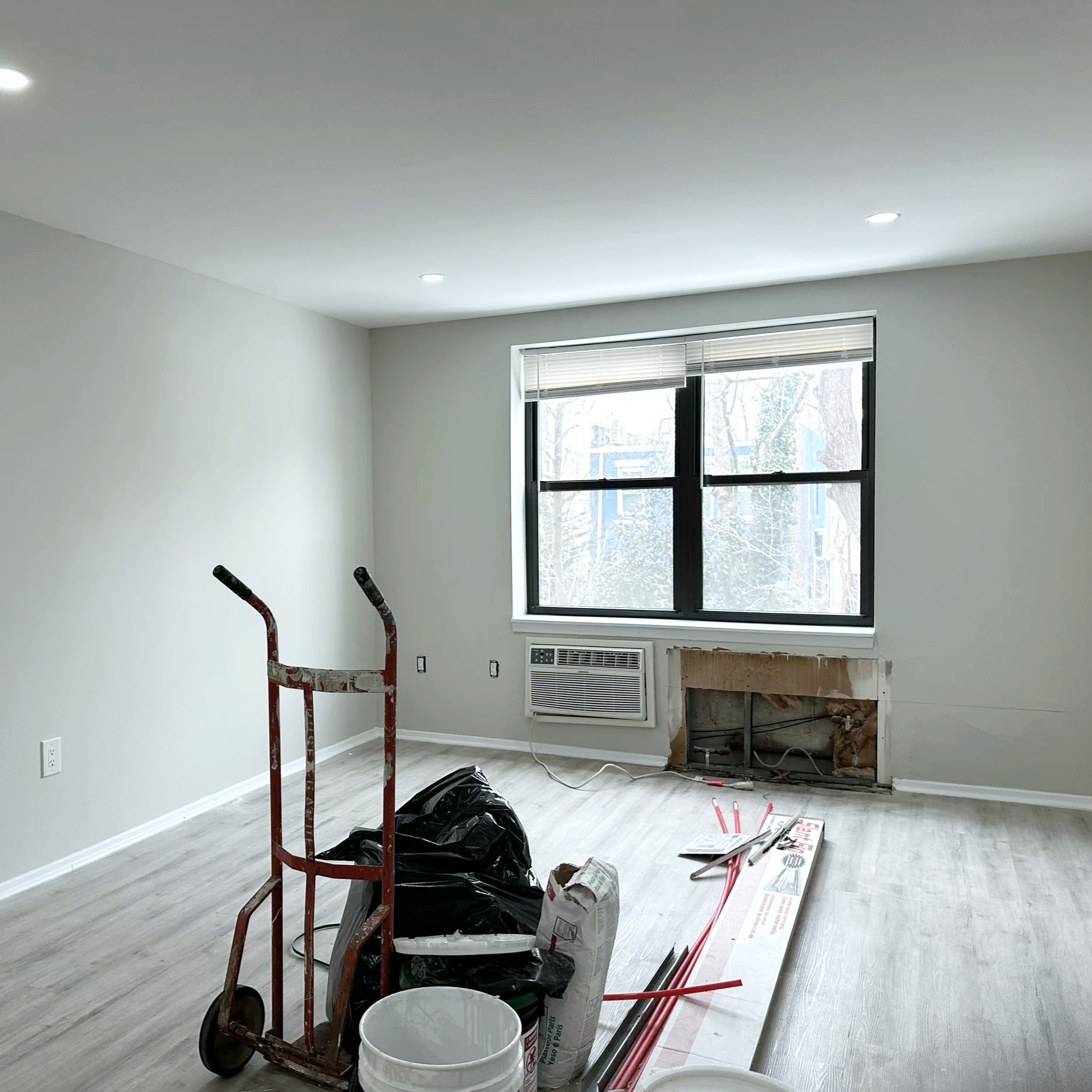 Empty room with ongoing renovation, equipment and materials including a red hand truck, plastic bags, pipes, and a window showing trees outside.