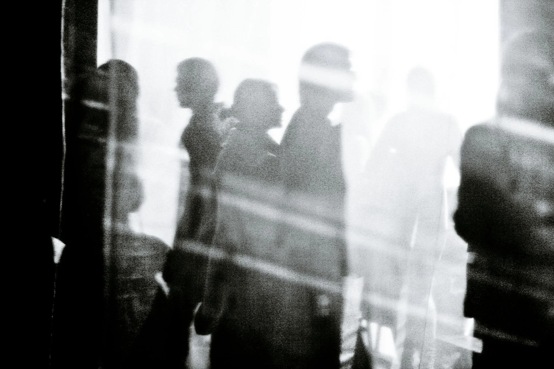 Silhouettes of people standing in bright light, with some wearing hats, in a blurred, black-and-white scene.