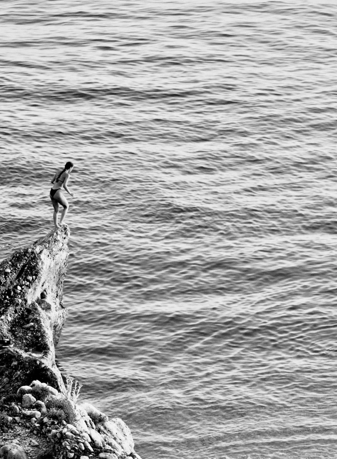 A person in a swimsuit standing on the edge of a rocky cliff by the water, preparing to jump or dive into the ocean.
