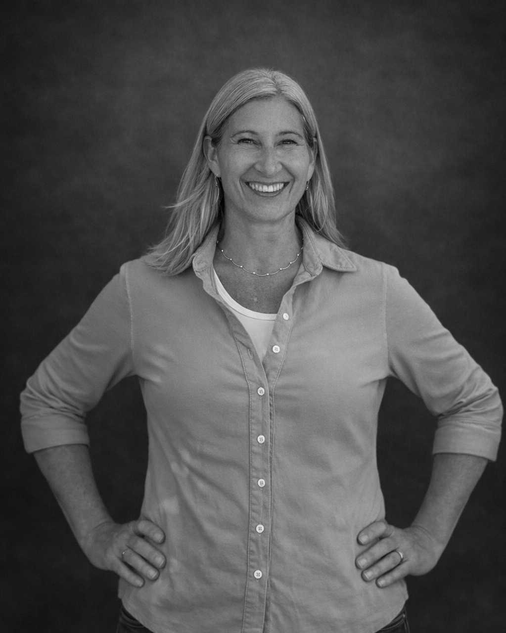 Smiling woman with shoulder-length light hair, wearing a button-up shirt and a necklace, standing with hands on hips against a plain dark background.