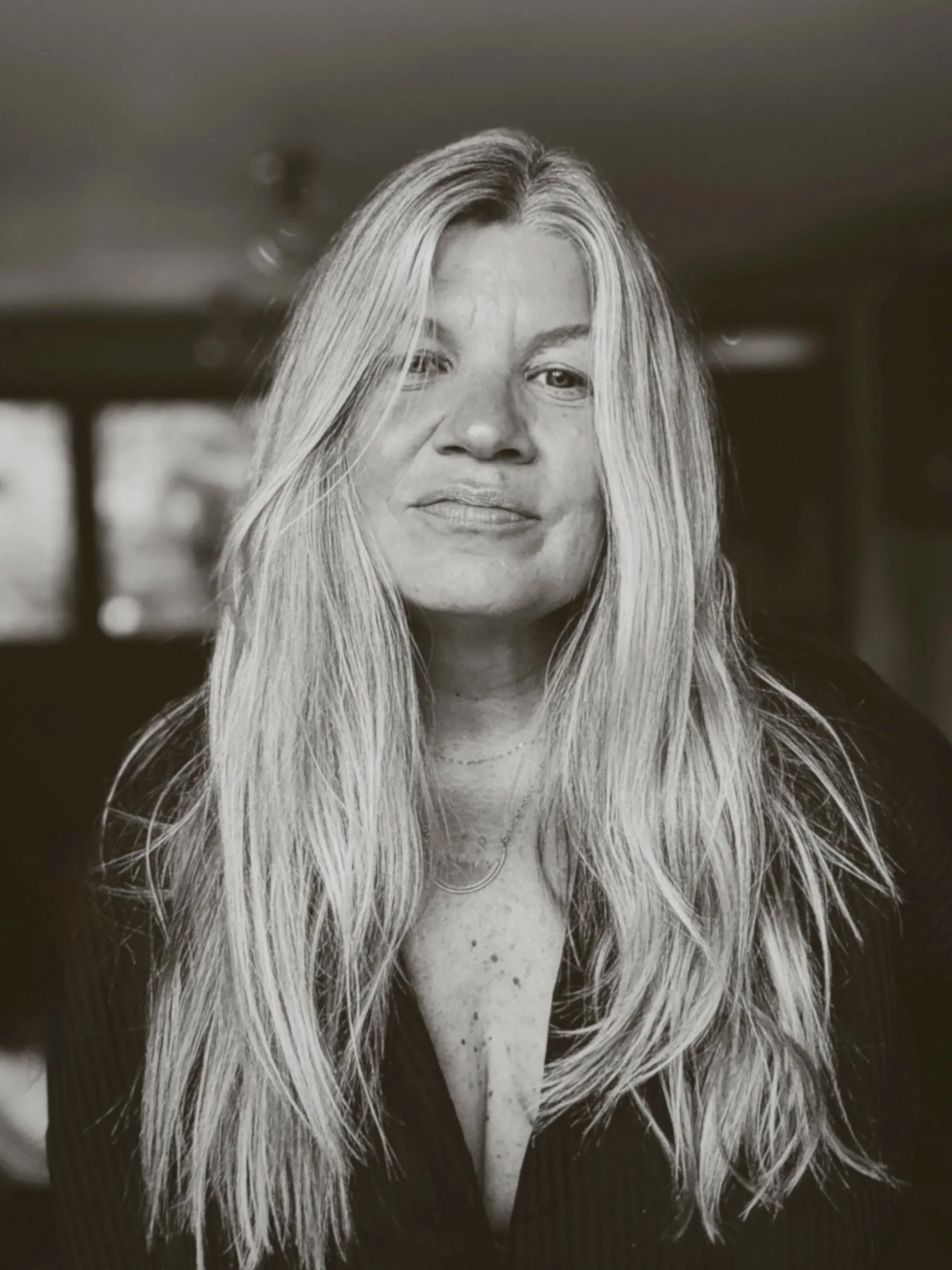 Black and white portrait of a woman with long, wavy blonde hair, wearing a dark top with a deep neckline, and looking directly at the camera.