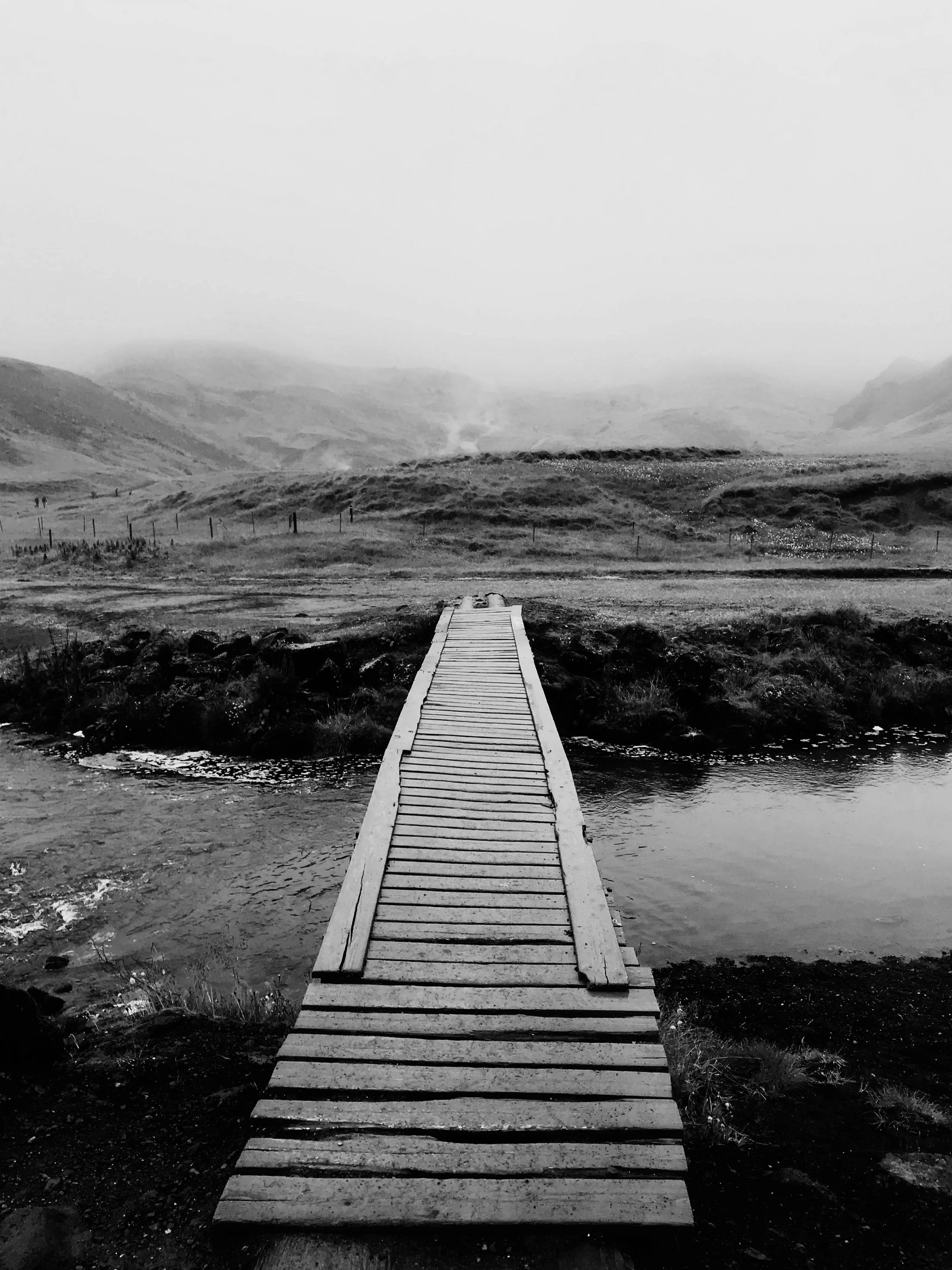 A wooden bridge extends over a small stream into a wide open landscape with rolling hills and mountains in the distance, under a cloudy sky.
