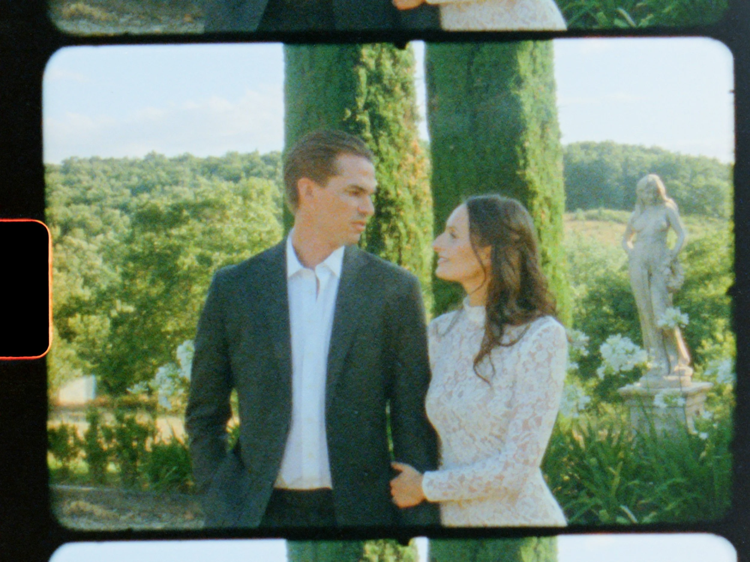 A man and woman are standing close together outdoors, looking into each other's eyes. The man is wearing a dark suit and white shirt, while the woman is dressed in a white lace dress. There are green trees and a statue of a woman in the background.