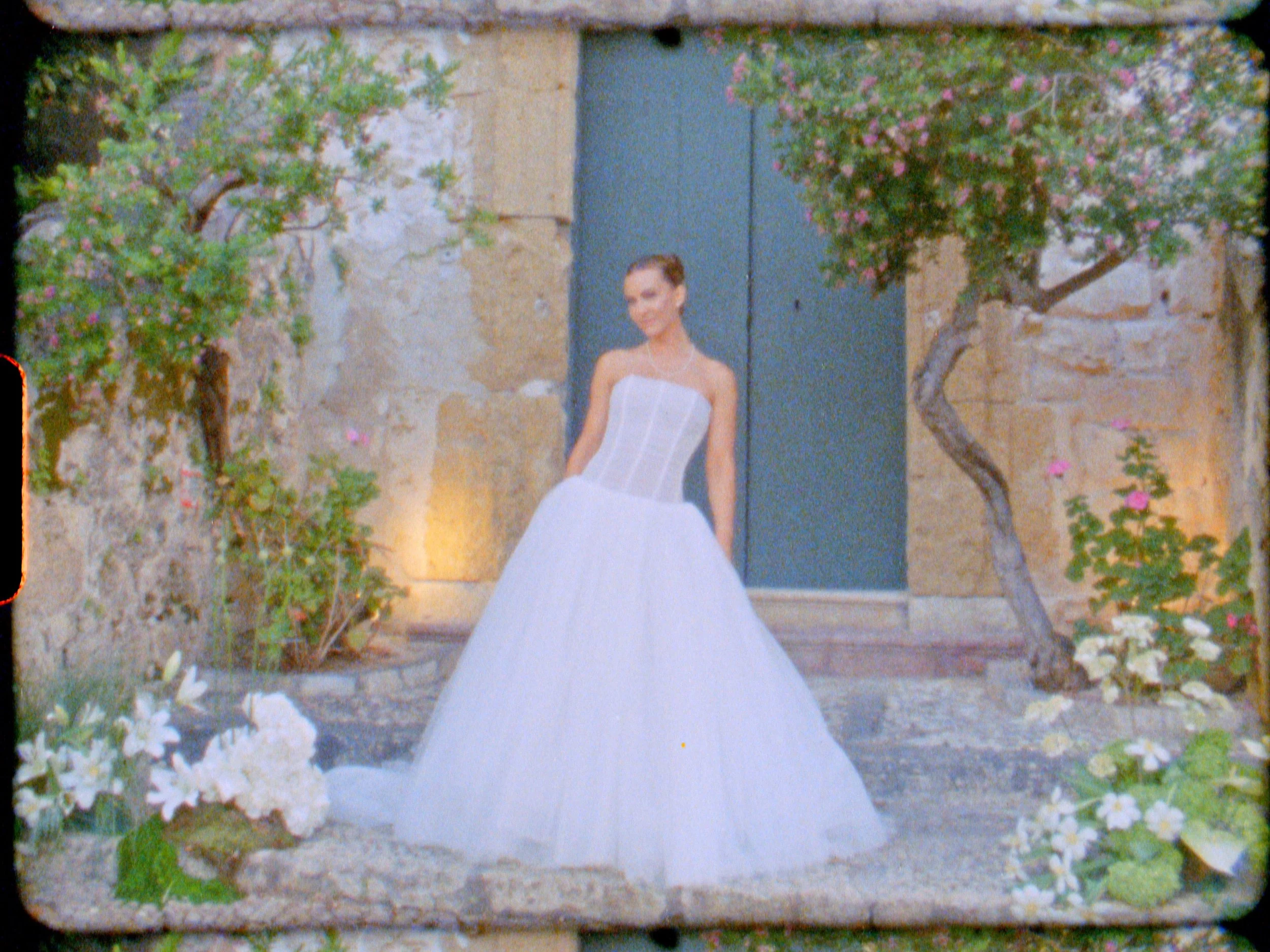 A woman in a white wedding dress standing in front of a stone wall with pink and white flowers and trees.