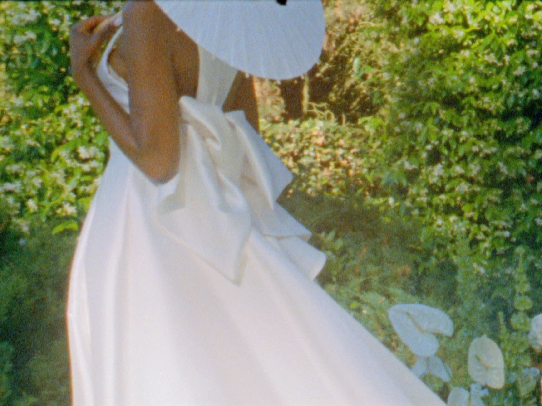 A woman in a white strapless dress holding a straw hat, standing outdoors among greenery and white flowers.