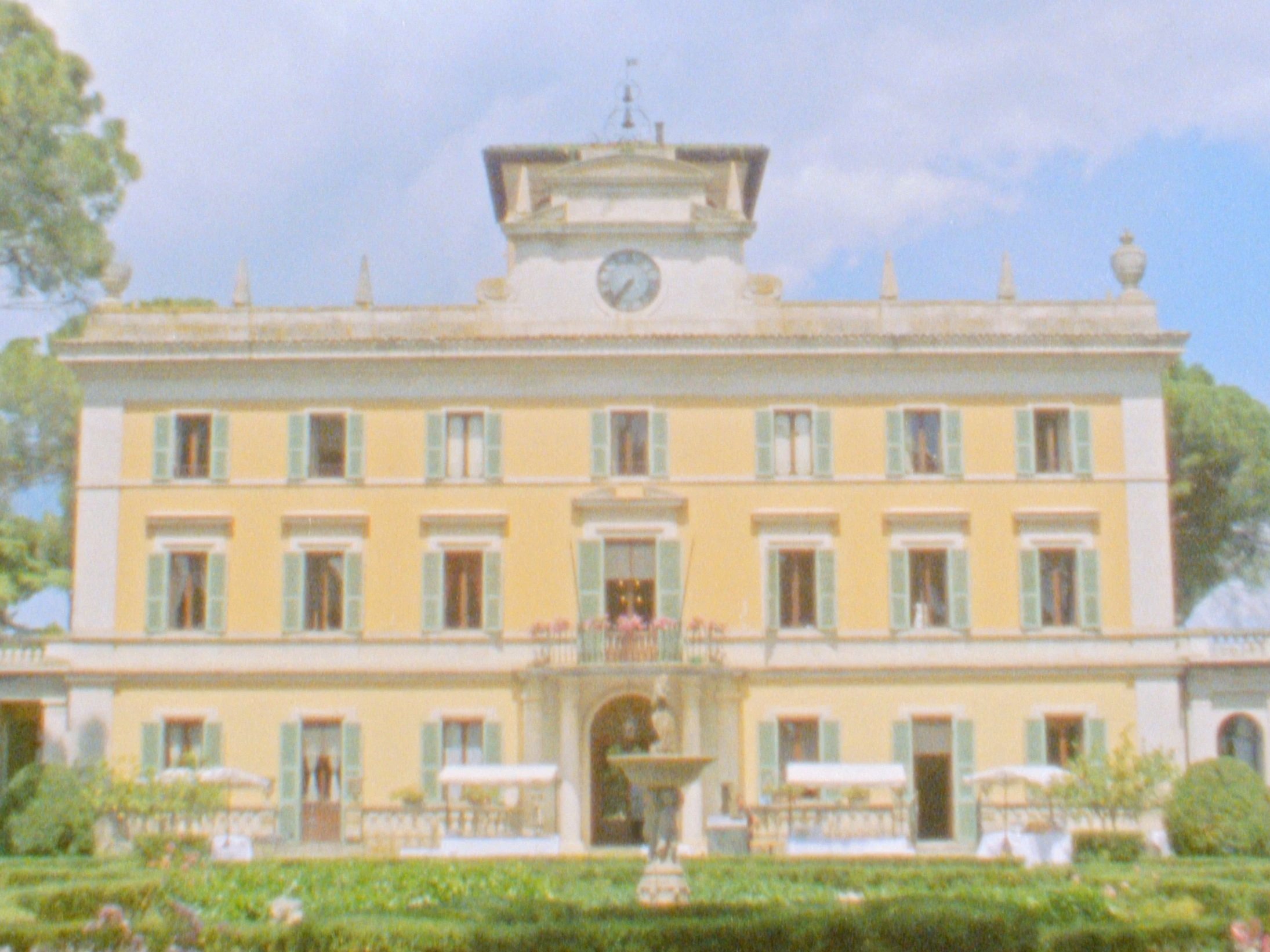 Yellow multi-story mansion with green shutters, fountain in front, surrounded by a garden, under a partly cloudy sky.