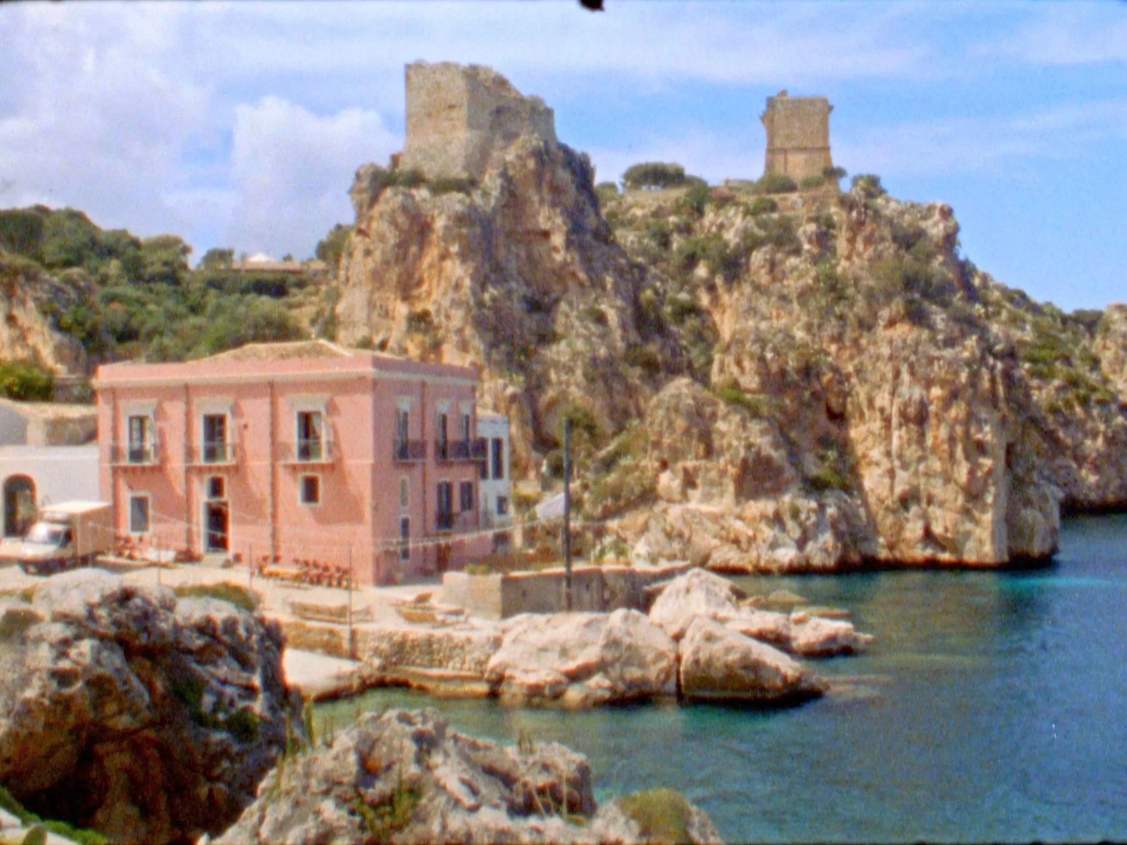 A pink building by a rocky coastline with a castle on a hill in the background.