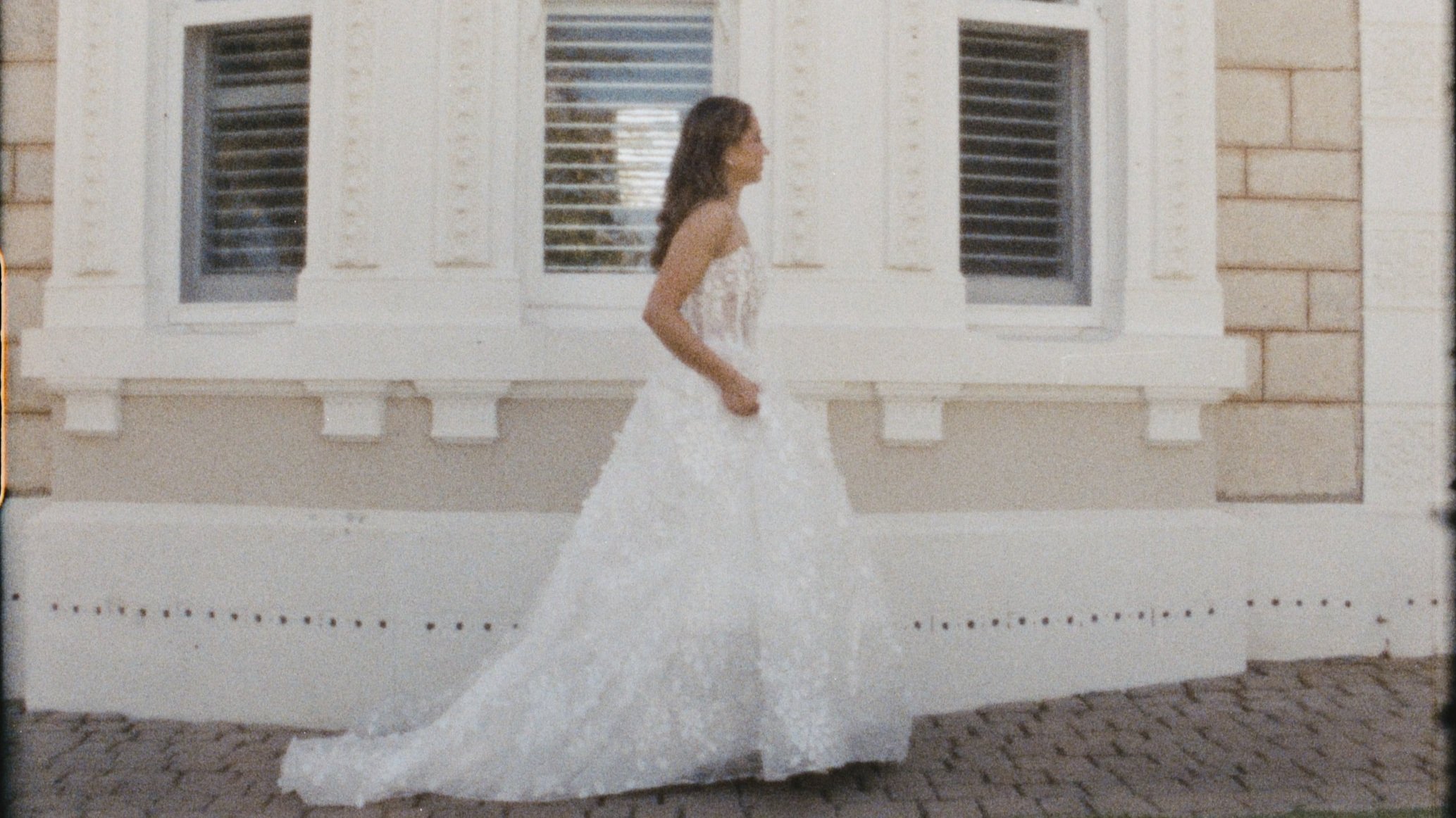 A woman in a white wedding dress walking past a building with cream brick and decorative window shutters.
