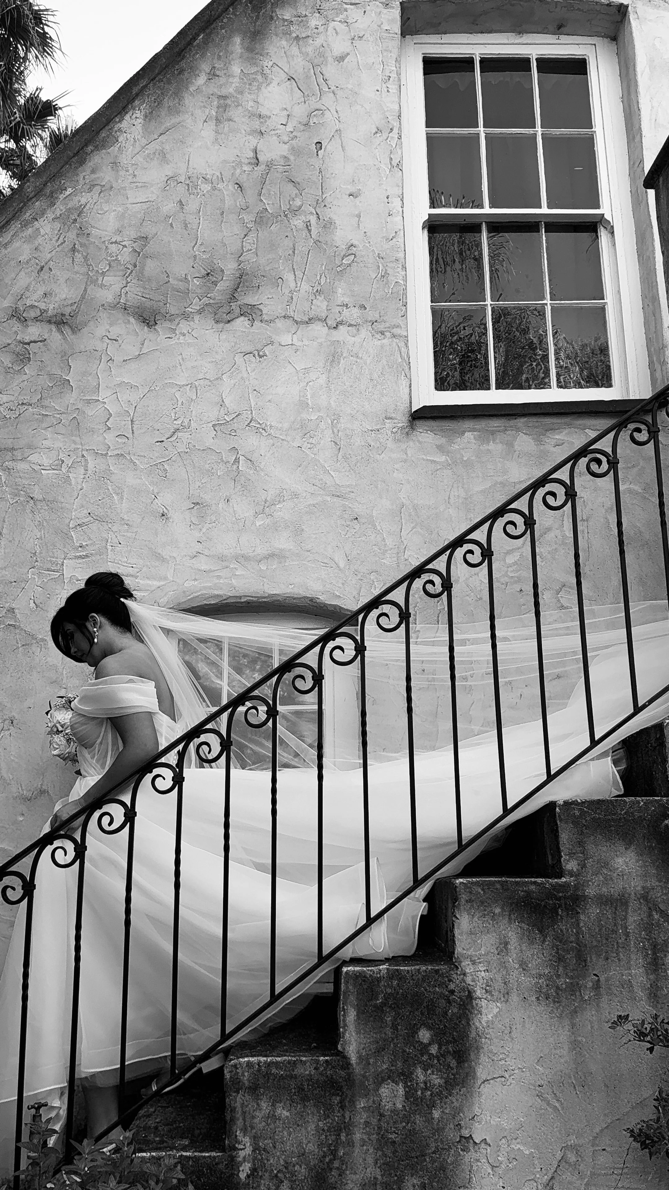 A bride in a wedding dress standing on outdoor stairs with a wrought iron railing and a large window on a textured wall in the background.
