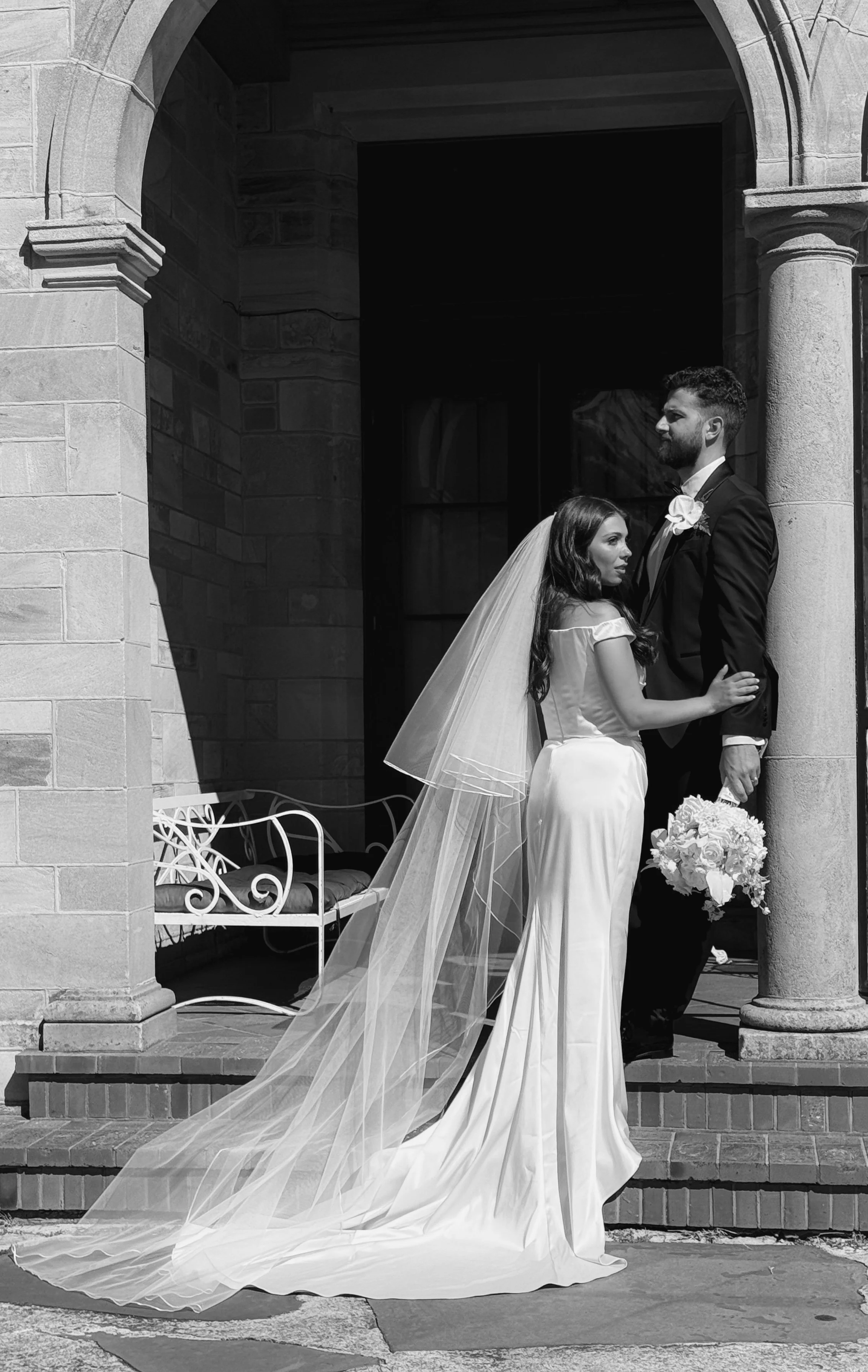 A bride and groom stand close together outside a church, with the bride wearing a long wedding gown and veil, and the groom in a tuxedo holding a bouquet of flowers, captured in black and white.